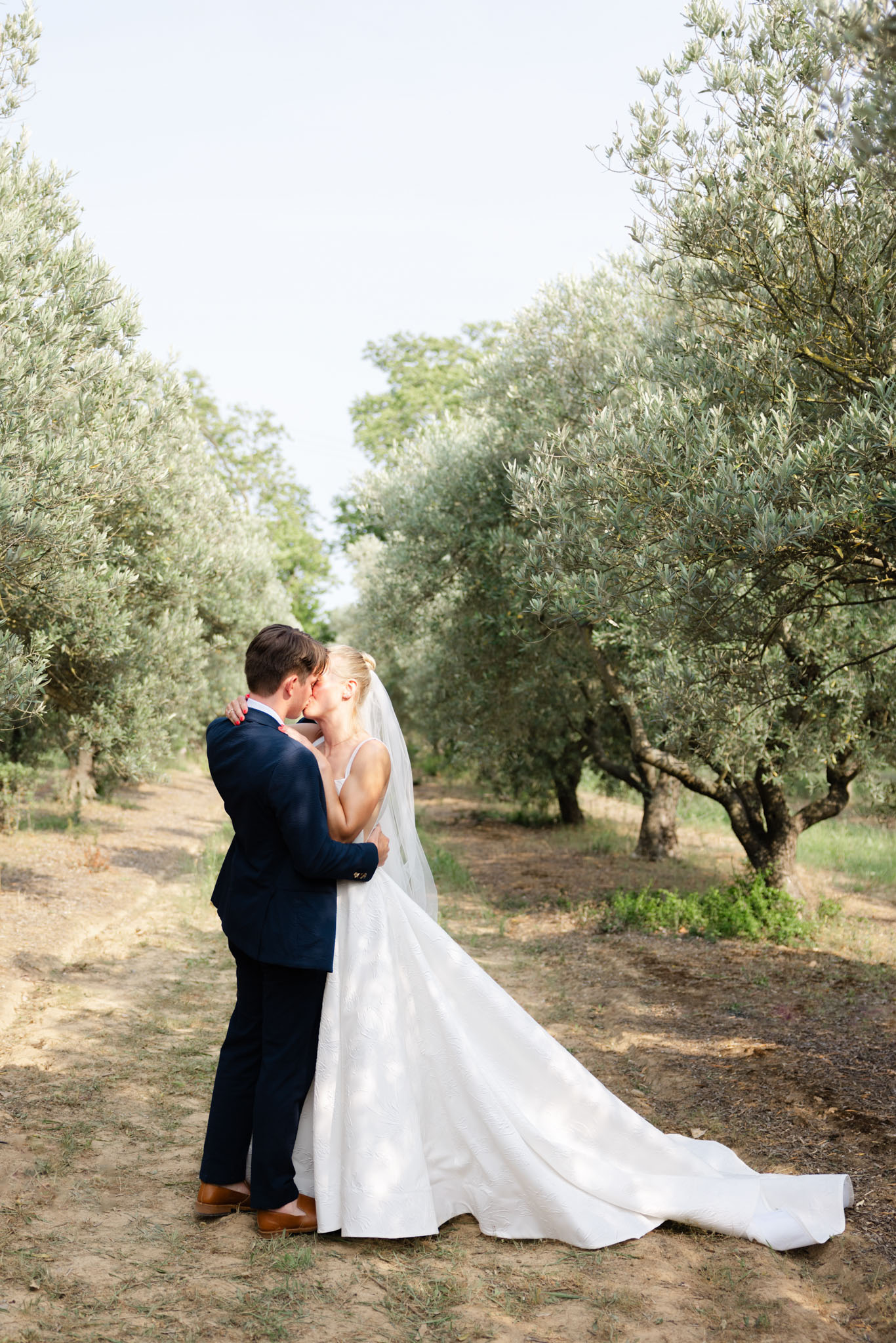 Bride and groom kiss on a dirt path through an olive grove, bride in white ballgown with cathedral veil