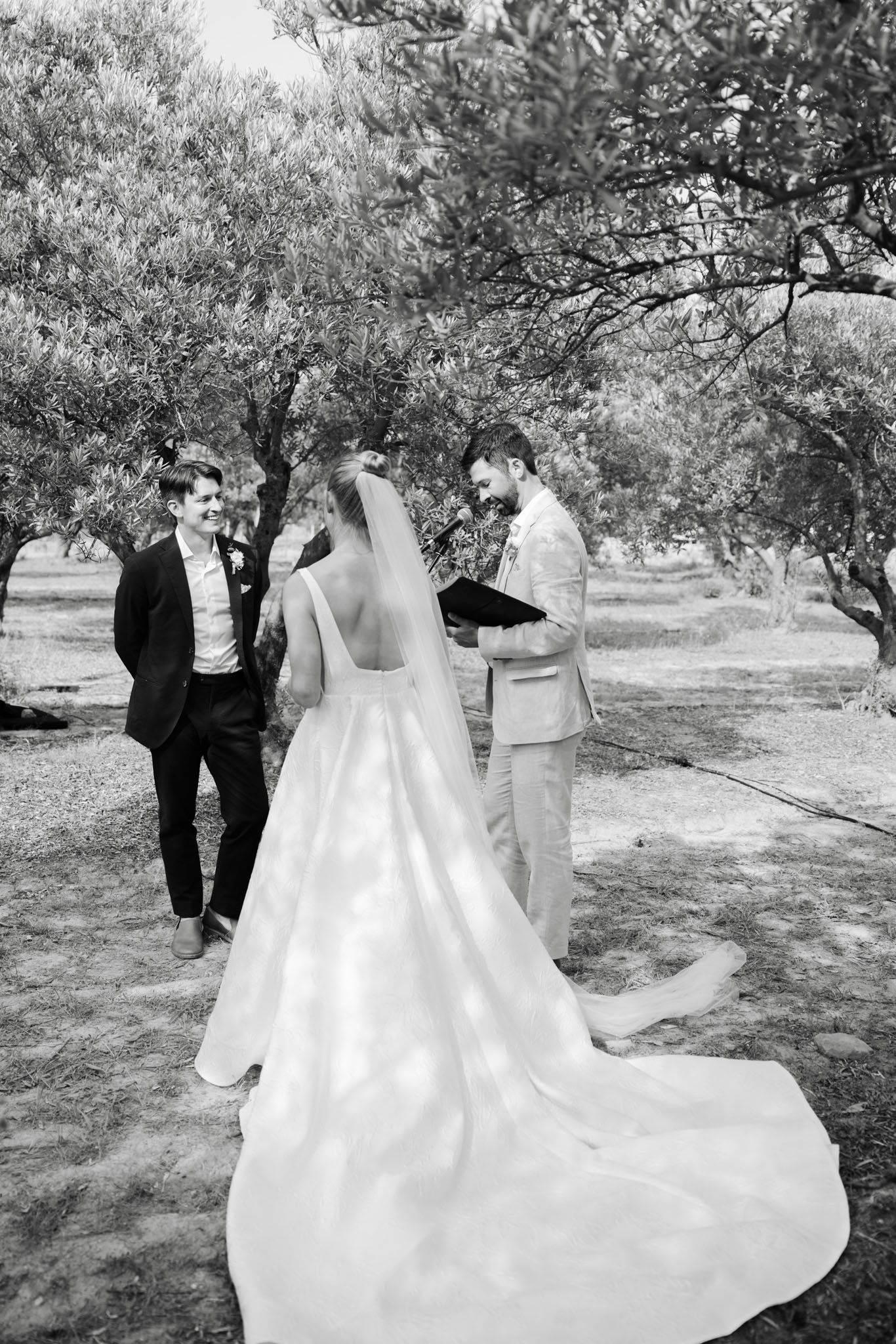 This black-and-white image captures an outdoor wedding ceremony taking place among a grove of olive trees. The bride is seen from behind, wearing a square-neck ball gown with a long cathedral train and a single-tier veil, her hair styled in an updo; she faces the groom, who wears a light-toned suit and holds an open ceremony booklet while speaking into a microphone. A second figure — likely the officiant or a groomsman — stands to the left in a dark suit with a boutonnière, smiling warmly toward the couple. The image is shot in high contrast black and white with bright highlights on the bride's dress and soft mid-tones throughout, and is framed as a medium wide shot taken slightly from above and behind the bride.
