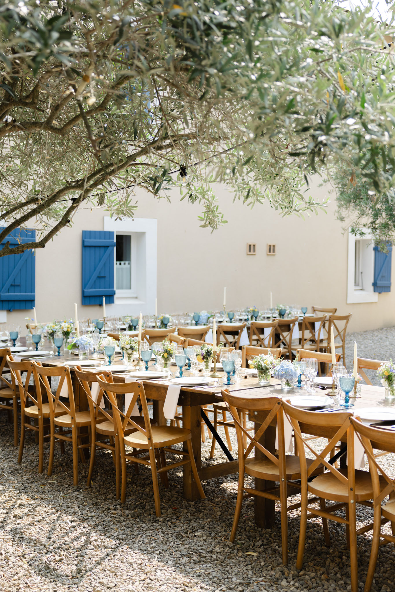 Farmhouse tables with blue tinted glassware hydrangea centerpieces brass candlesticks before Provencal building with cobal...