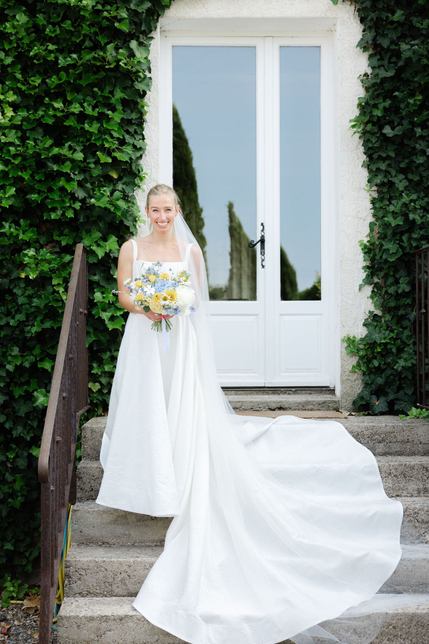 A bridal portrait taken outdoors on the stone steps of what appears to be a French country property, with a white-painted double door framed by ivy-covered walls serving as the backdrop. The bride stands alone, smiling directly at the camera, wearing a white square-neck gown with thick straps, a textured fabric skirt with a long cathedral train, and a sheer veil. She holds a round bouquet featuring yellow ranunculus, white daisies, pale blue hydrangea, and blue nigella flowers. The composition is a full-length portrait shot, capturing the full spread of the train across the steps.