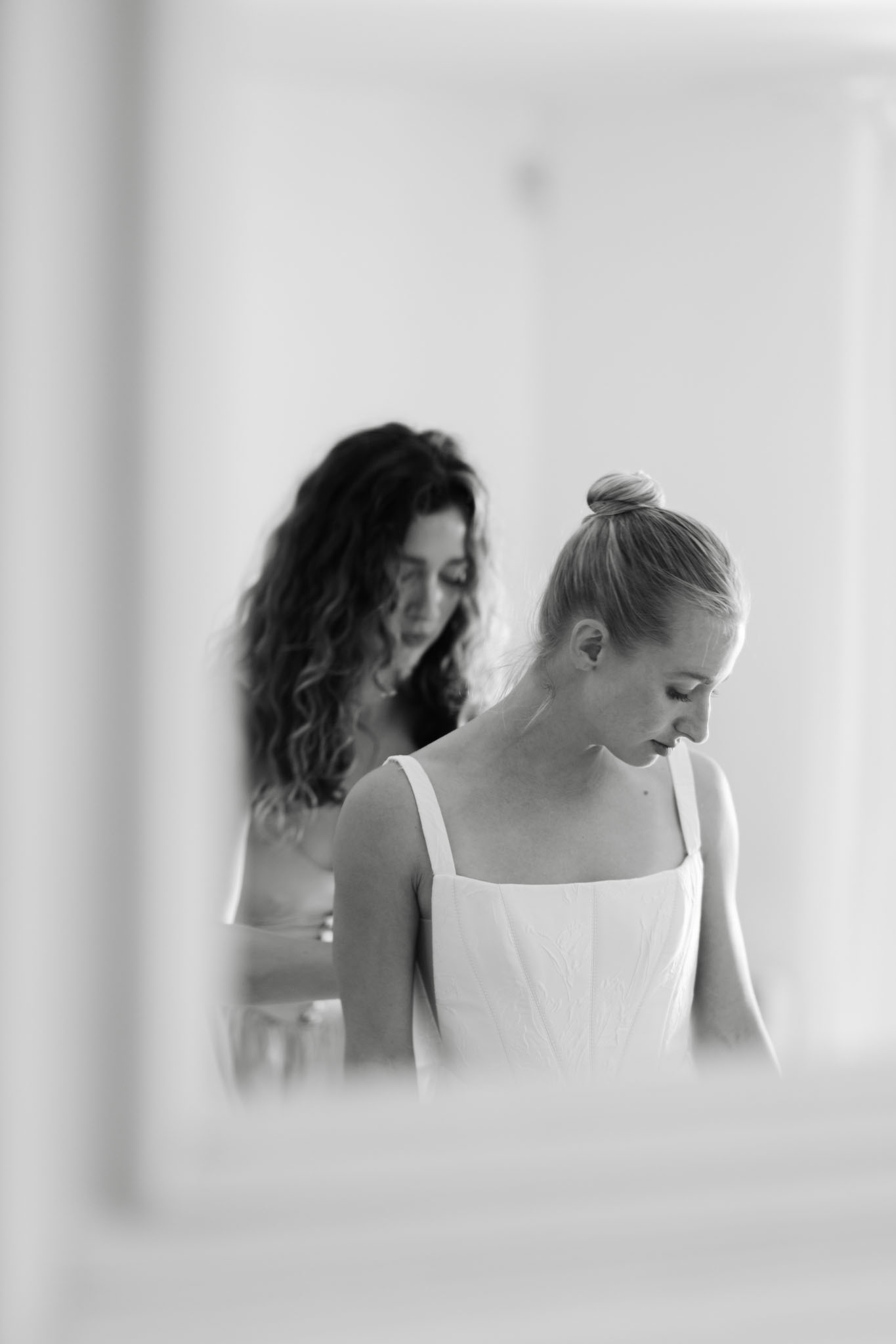 A black-and-white getting-ready scene showing the bride in a structured white wedding dress with square neckline and wide straps, her blonde hair pulled into a high bun, looking downward as a second woman with long dark curly hair fastens or adjusts the back of the gown. The setting is a bright, light-toned indoor room with white walls. The image is framed through a soft foreground blur, likely a doorframe or mirror edge, giving depth to the composition. The overall tones are high-key with soft contrast, and the styling appears clean and modern.