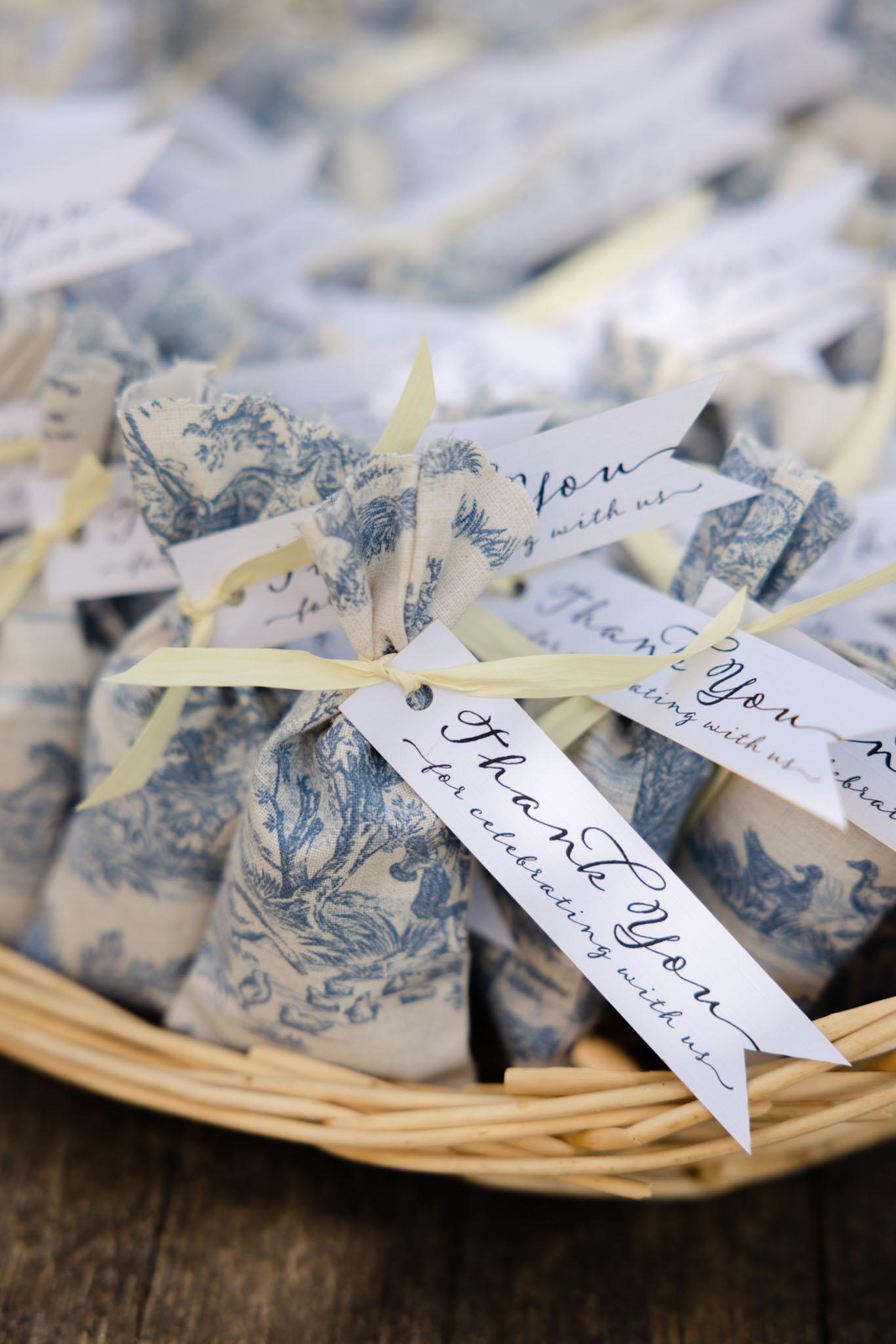 Close-up detail shot of wedding guest favors displayed in a wicker basket on a wooden surface. Each favor is a small pouch or wrapped item made from blue and cream toile de Jouy fabric, tied with a pale yellow satin ribbon and finished with a white calligraphy tag reading 'Thank You for celebrating with us.' The basket contains numerous identical favors, with the background ones softly blurred. The styling palette of blue toile, cream, and pale yellow points to a classic French-inspired aesthetic.