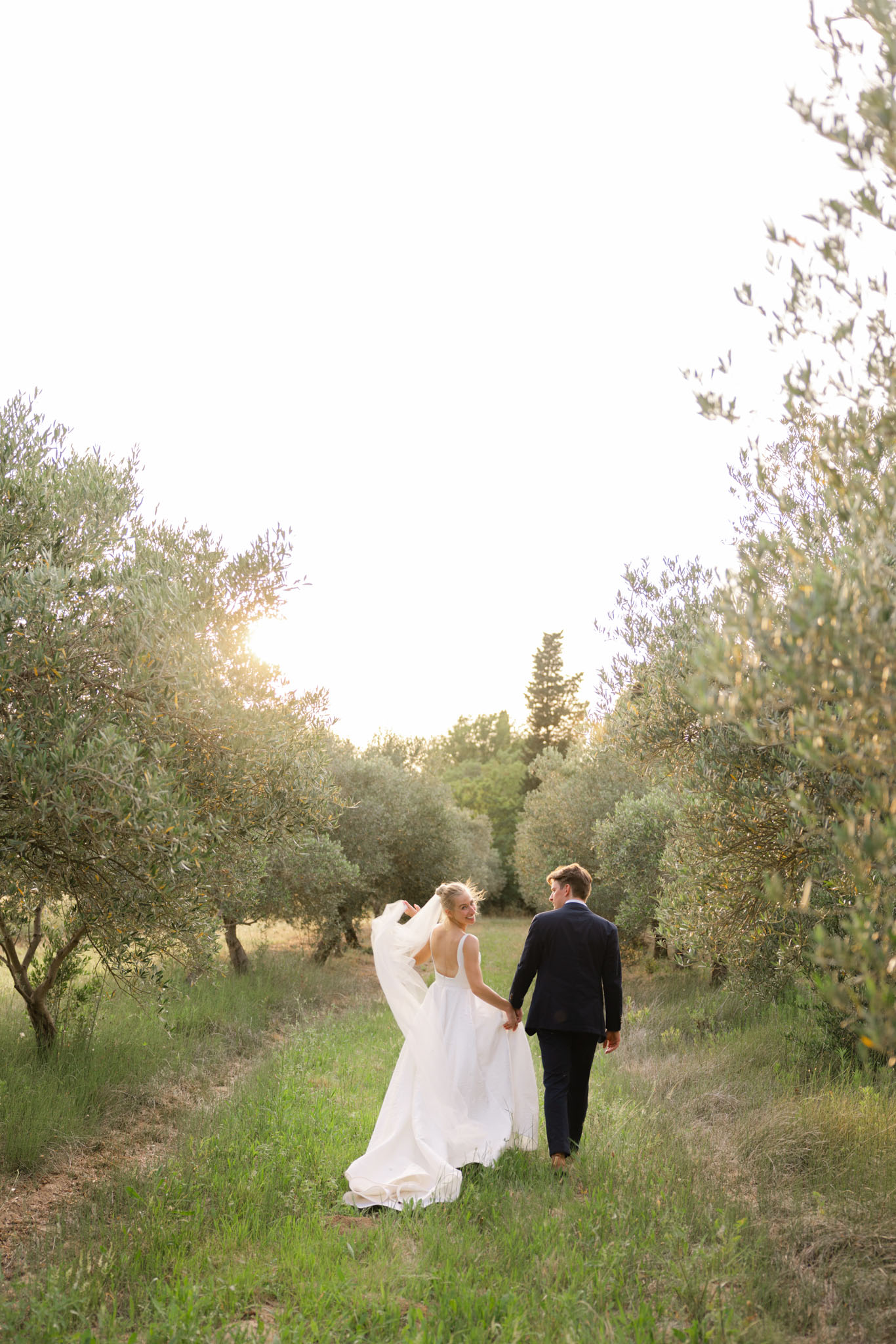 A couple portrait taken outdoors during golden hour, with the bride and groom walking hand-in-hand along a grass path between two rows of olive trees. The bride, wearing a white full-skirted gown with an open back and a long flowing veil, turns back to look over her shoulder with a smile. The groom is dressed in a navy suit. The image is a wide shot taken from behind, with warm backlit sunshine creating a soft glow through the olive grove. The overall style is natural and relaxed, consistent with a South of France or Provençal outdoor setting.