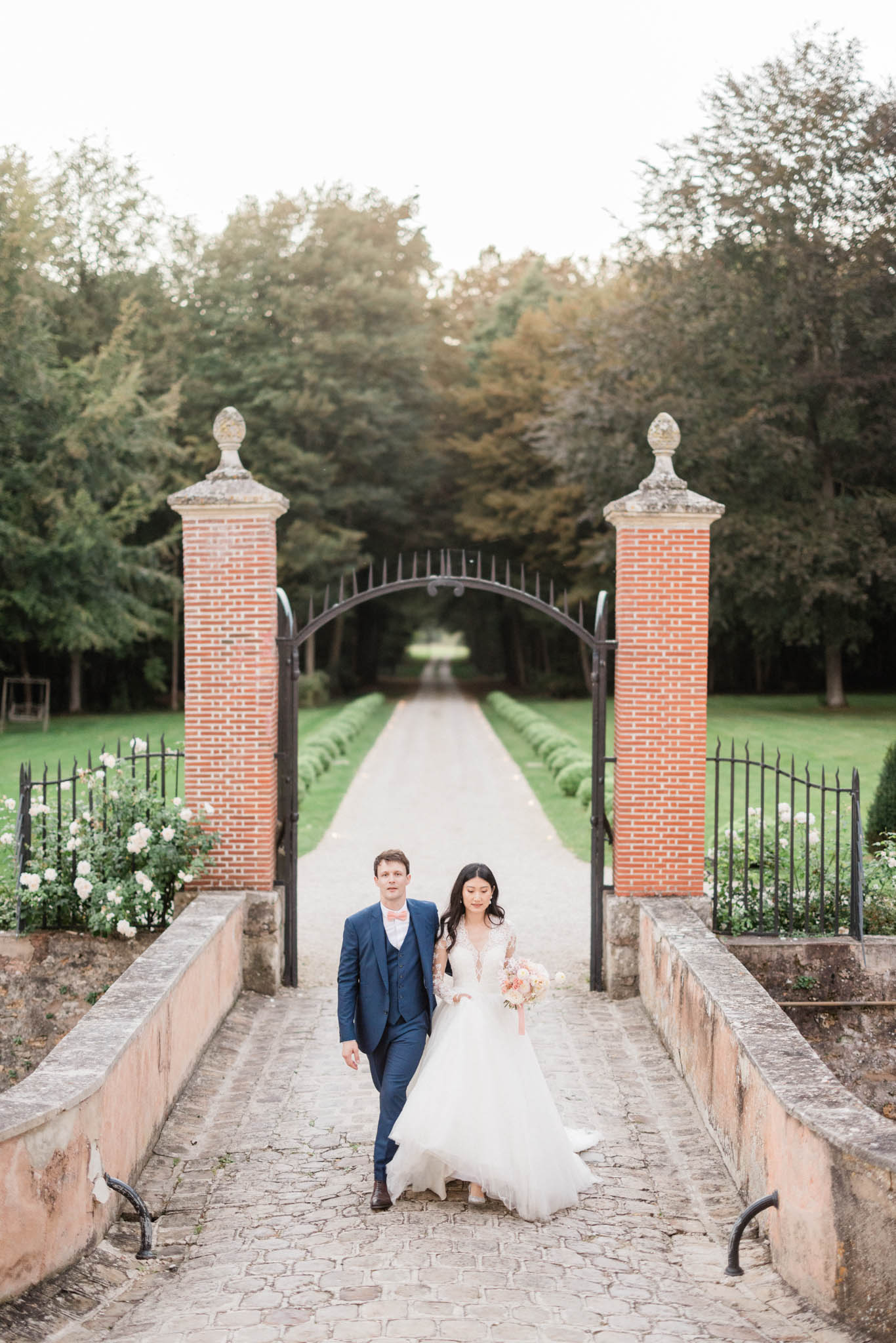 A couple portrait taken outdoors at what appears to be a French château or estate, with the bride and groom walking together down a cobblestone path toward the camera. The groom wears a navy three-piece suit with a pink bow tie and brown dress shoes, while the bride wears a white long-sleeved lace and tulle ballgown with a deep V-neckline; she carries a bouquet featuring blush, peach, and ivory blooms. Behind them, a formal wrought-iron gate flanked by red brick pillars with stone finials frames a long, symmetrical tree-lined allée that recedes into the distance, with white rose bushes visible on either side of the gate. The composition is a centered medium-wide portrait shot with the gate and allée serving as a strong leading-line backdrop, giving the image a classic, formal French estate aesthetic.