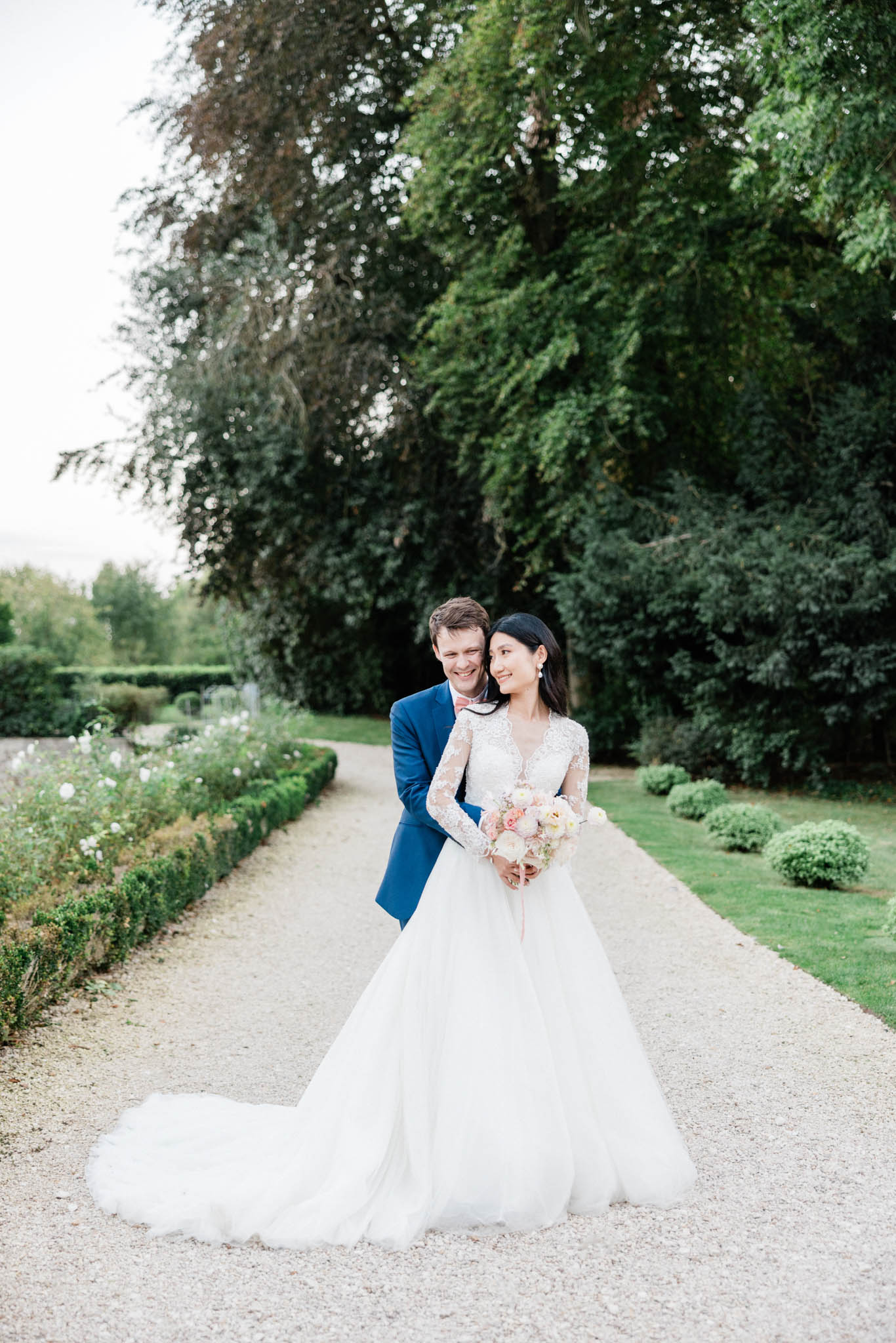 Bride in white lace-sleeve ball gown and groom in navy suit on gravel path in formal French garden