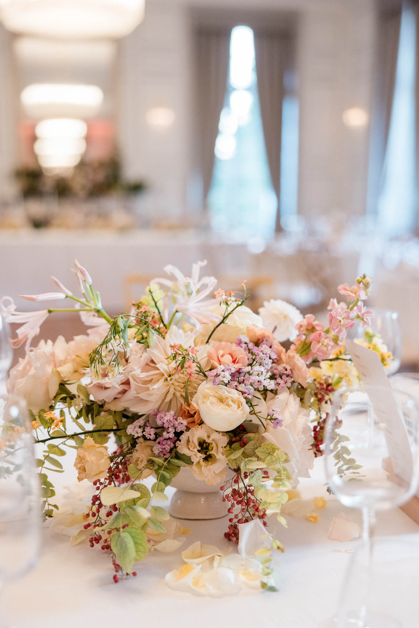 Close-up detail shot of a low floral centerpiece arranged in a cream ceramic pedestal vase, placed on a white linen-covered reception table inside a bright ballroom or formal indoor venue. The arrangement includes cream garden roses, blush dahlias, peach spray roses, pale pink larkspur, mauve wax flower, yellow lisianthus, trailing eucalyptus, and cascading red pepper berries, with scattered petals falling onto the tablecloth. The overall floral palette is soft and romantic, mixing creamy whites, blush, peach, and pale pink tones with touches of deep red from the berries. Clear glassware is partially visible to the right, and the blurred background reveals additional table arrangements, overhead lighting, and tall windows with sheer curtains in a light-filled reception room.