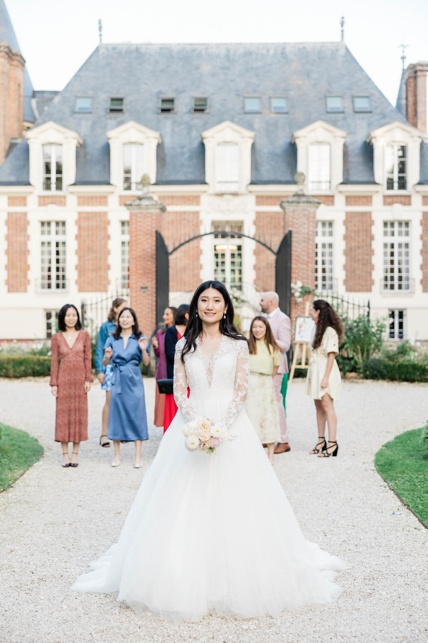 A bride stands in the foreground on a gravel courtyard in front of a French château, facing the camera and smiling. She wears a white ball gown with long lace illusion sleeves and a full tulle skirt, and holds a bouquet of blush pink and ivory blooms. Behind her, approximately seven guests in colorful attire — including a dusty blue satin wrap dress, a terracotta midi dress, a red dress, a pale yellow dress, and a pink blazer — are gathered and socializing near the château's black iron gate entrance. The setting is an outdoor courtyard of a classic French brick-and-stone château with slate mansard roofing and white-trimmed dormer windows. The shot is a medium-wide portrait with the bride sharply in focus and the guests and building softly blurred in the background, suggesting a candid moment during cocktail hour or between portrait sessions. Potential venue feature image.