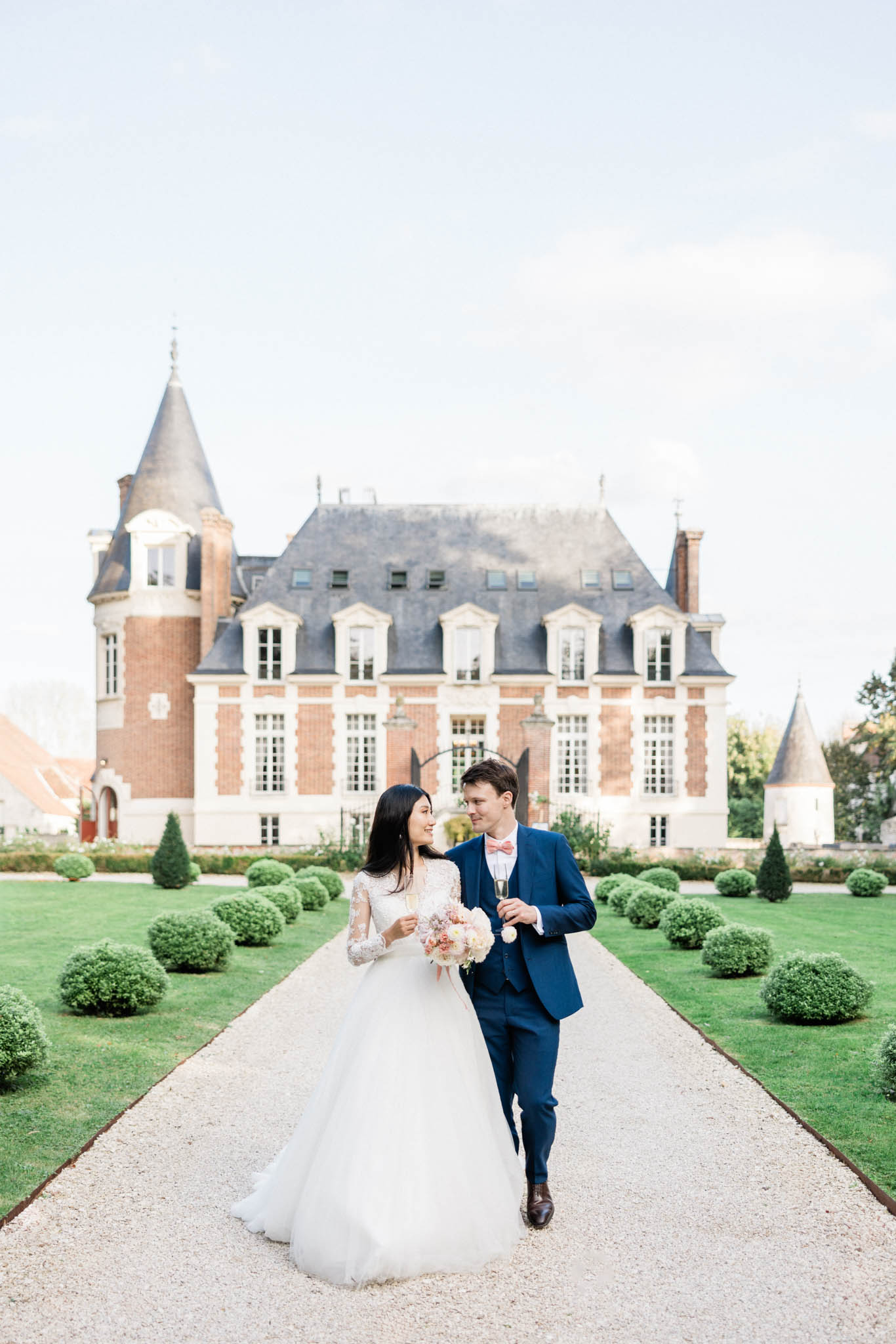 Bride in lace ballgown with blush bouquet and groom in navy suit walking from red-brick chateau with champagne