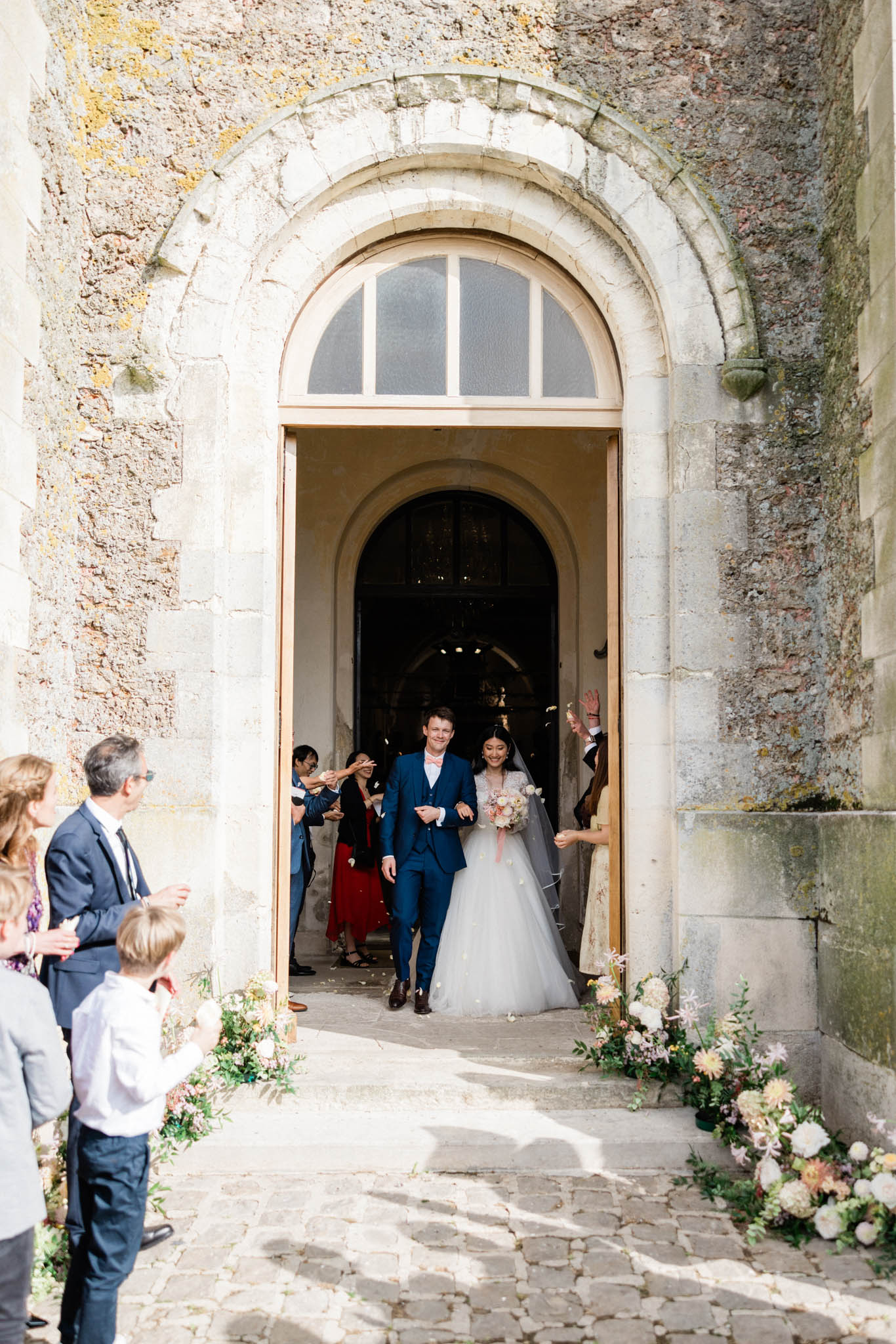 The couple is exiting a stone chapel through a large arched doorway after their ceremony, walking out together as guests on either side applaud and toss confetti. The groom wears a navy blue three-piece suit with a pink bow tie, and the bride wears a full ballgown with a tulle skirt, floral appliqué bodice, and a long veil, carrying a loose bouquet of blush, peach, and ivory blooms. The chapel entrance is flanked by low floral arrangements on the cobblestone steps featuring white and blush dahlias, peach blooms, and trailing greenery in a garden-style arrangement. Approximately 10–15 guests are visible in the background and to the sides, with the chapel's arched stone doorway and interior arch visible behind the couple in this medium wide shot.