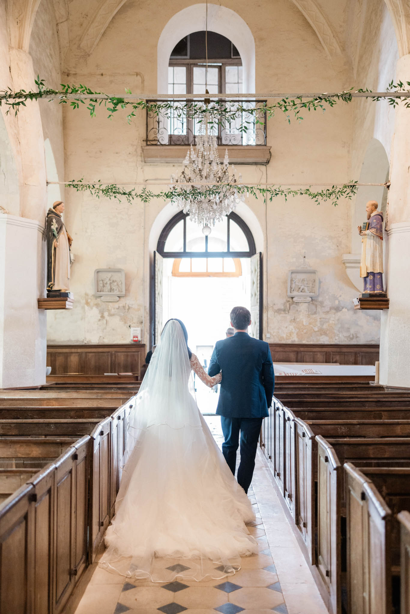 Couple walks down chapel aisle from behind with green garlands strung across nave and geometric tile floor