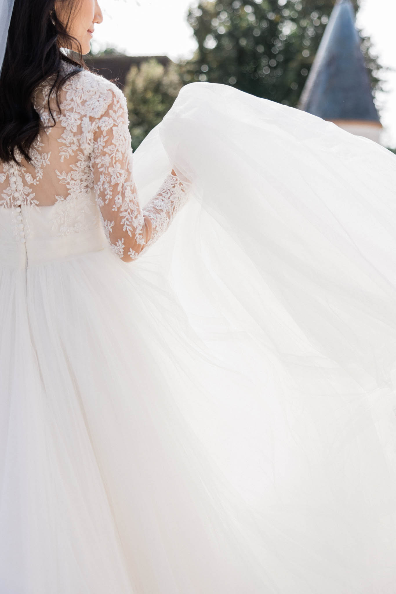 A close-up portrait of a bride shot from behind, outdoors, with a château turret visible in the background. She is wearing an ivory ball gown with a sheer, illusion long-sleeved bodice featuring intricate floral lace appliqué, and a full voluminous tulle skirt that fans out dramatically to fill the lower half of the frame. The bride, with dark wavy hair, is posed in three-quarter profile looking to one side while holding the skirt of her dress. The composition emphasizes the contrast between the delicate lace detailing on the back of the bodice and the soft, sweeping volume of the tulle skirt, shot in soft natural daylight.