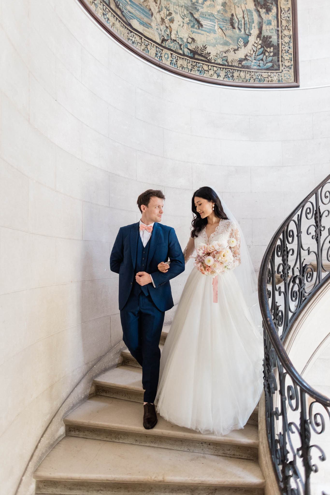 A couple portrait taken on a curved stone staircase inside what appears to be a French château, shot from a slightly elevated angle. The groom wears a navy three-piece suit with a coral-pink bow tie and brown dress shoes, while the bride wears a full-length ivory ballgown with a lace long-sleeve bodice, a tulle skirt, and a cathedral-length veil. She carries a loose bouquet of cream ranunculus, blush dahlias, and dried pampas-style elements tied with a dusty pink ribbon. A large antique tapestry with blue and green tones is visible on the curved wall above them, and a wrought-iron spiral railing frames the right side of the frame. The overall styling reads as classic French château with a soft blush and navy color palette.