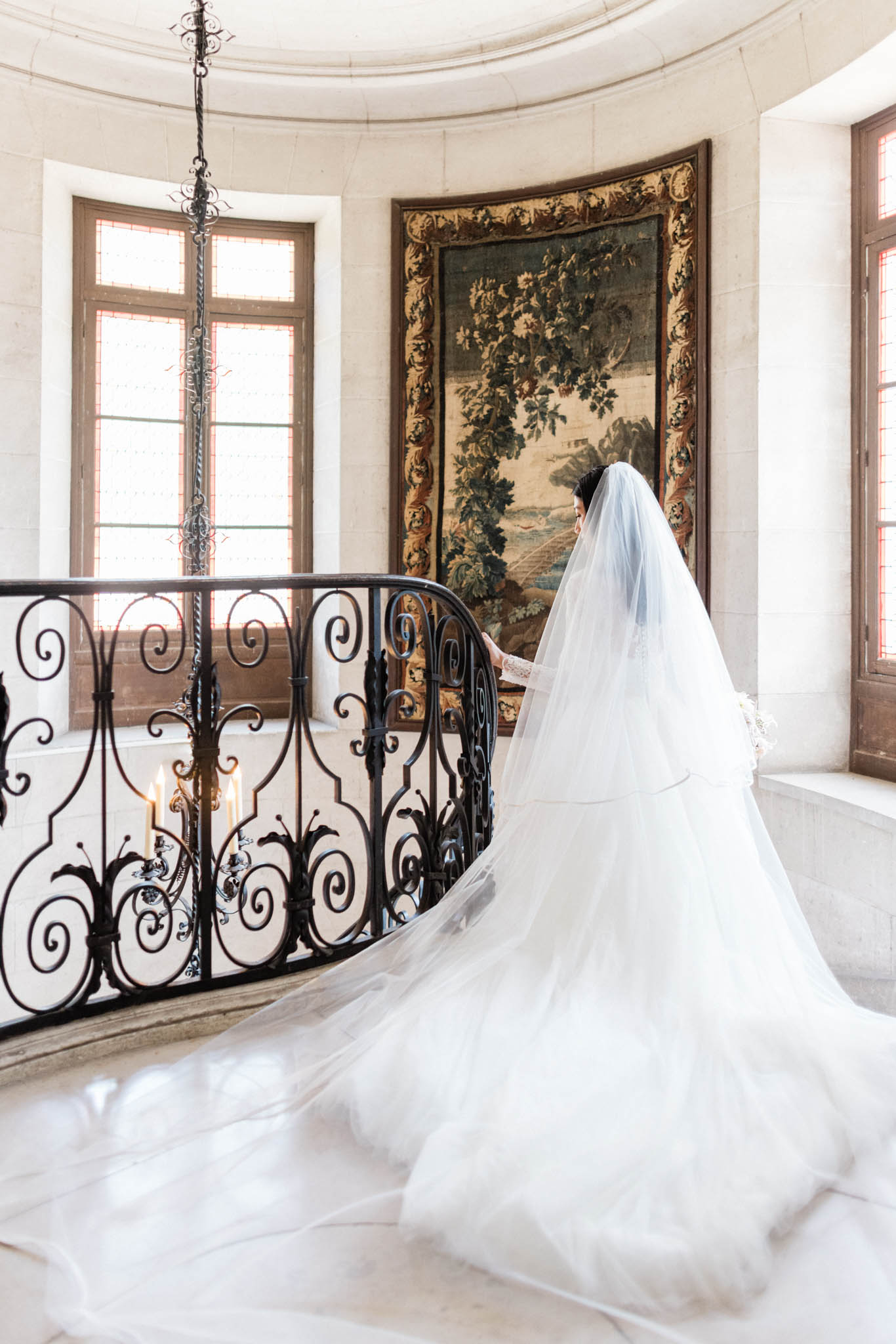 A bridal portrait taken indoors in what appears to be a French château, showing the bride from behind as she stands at the top of a curved staircase with ornate black wrought-iron balustrades. The bride wears a full-skirted white ball gown with an exceptionally long cathedral-length tulle veil that pools dramatically across the marble floor. She appears to be reading from a small booklet or card. The circular landing features tall arched windows with warm natural light, a large gilded-framed tapestry depicting a landscape and foliage scene on the wall behind her, and a candelabra with lit candles mounted to the ironwork railing. The composition is a wide-to-medium portrait shot from a slightly elevated angle, emphasizing the volume and spread of the dress and veil against the light marble floor. Potential venue feature image.
