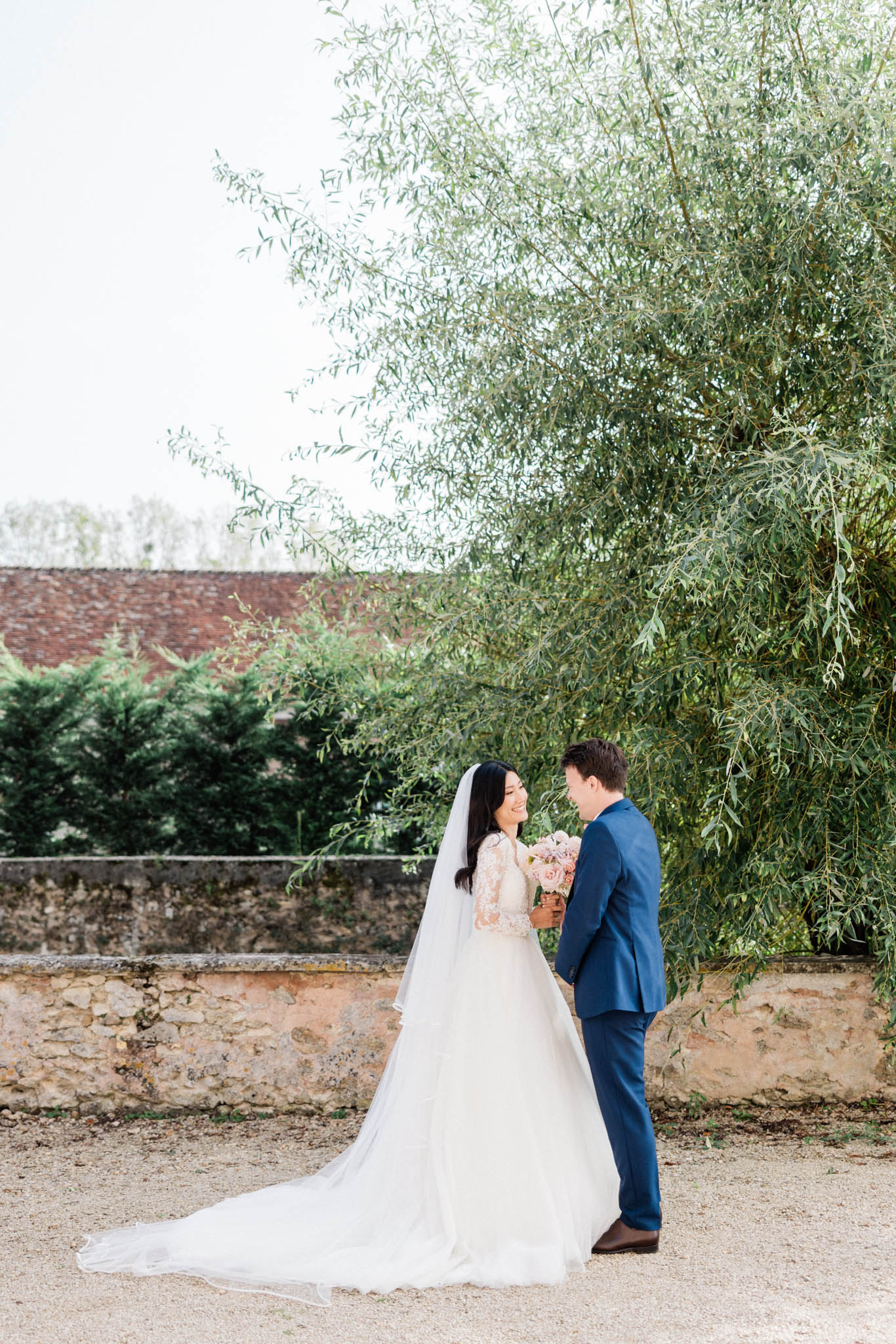 A couple portrait taken outdoors on a gravel courtyard, with the bride and groom facing each other and smiling. The bride wears a white ballgown with a lace long-sleeve bodice and a cathedral-length veil, and holds a blush pink bouquet of roses. The groom wears a navy blue suit with no tie. The setting appears to be a French country estate or château grounds, with a rustic stone boundary wall and a terracotta-roofed outbuilding visible in the background. The shot is a full-length portrait with a soft, airy feel in natural daylight.