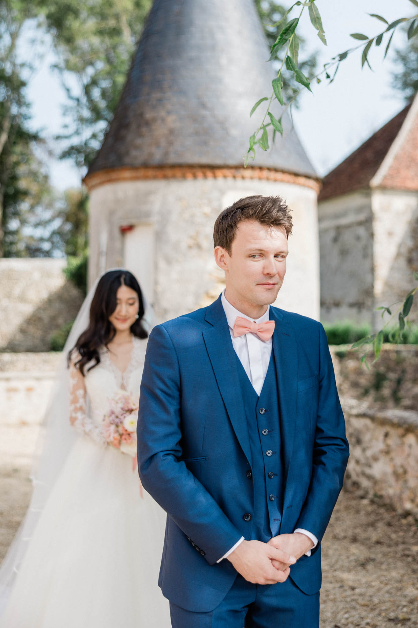A first-look moment captured outdoors at a French château, with the groom standing in the foreground facing away from the bride as she approaches from behind. The groom wears a navy blue three-piece suit with a salmon-pink bow tie and white dress shirt, hands clasped in front of him. The bride wears a white gown with lace-embroidered long sleeves and a deep V-neckline, carrying a loose bouquet in blush, peach, and lilac tones; her long dark hair falls in waves over her shoulders. The background features the rounded stone tower of a château with a conical slate roof, consistent with classic French architecture. The composition is a medium portrait shot with the groom sharply in focus in the foreground and the bride softly blurred in the mid-ground, creating depth.