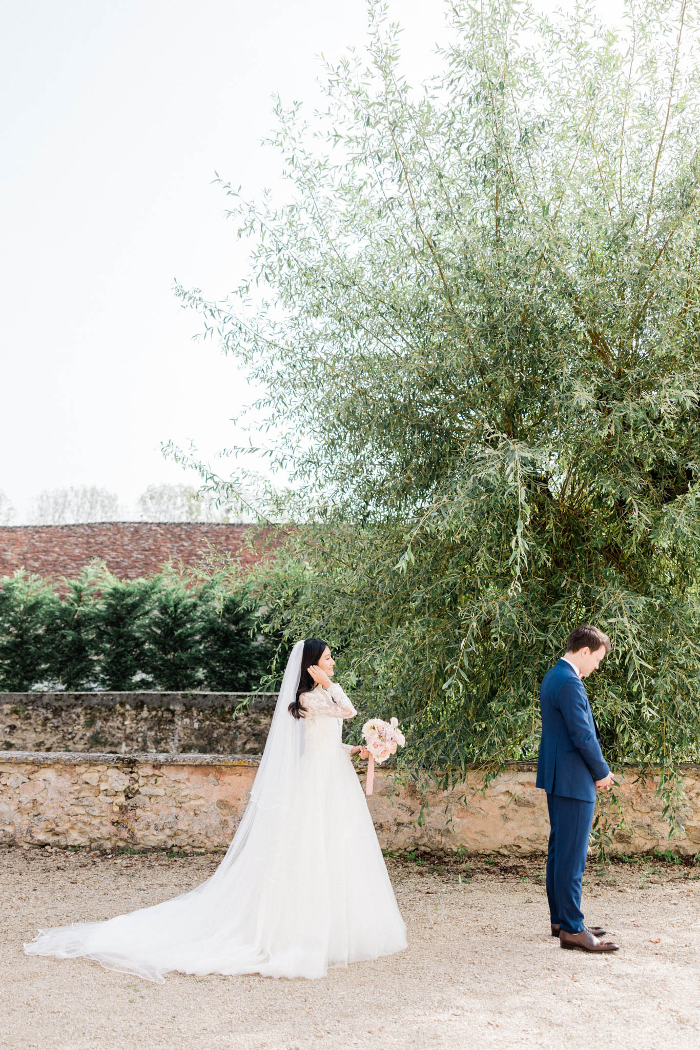 A first look moment captured outdoors on a gravel courtyard, with the bride and groom standing apart and facing away from each other in anticipation. The bride wears a long-sleeved, lace-detailed ivory ballgown with a full tulle skirt and a long cathedral-length veil, and holds a blush pink bouquet of what appear to be roses and peonies; she raises one hand to her face in an emotional gesture. The groom wears a navy blue suit with brown leather dress shoes and stands with his head bowed, hands clasped behind his back. The wide-format portrait is shot in a classic, airy style with soft natural light against a backdrop of an old stone perimeter wall with a terracotta-tiled roof visible beyond it.