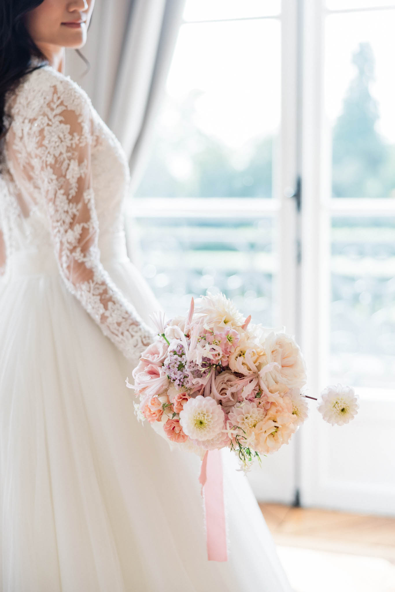 Bride in long-sleeve lace gown holding a blush and mauve bouquet by tall French doors with soft natural light
