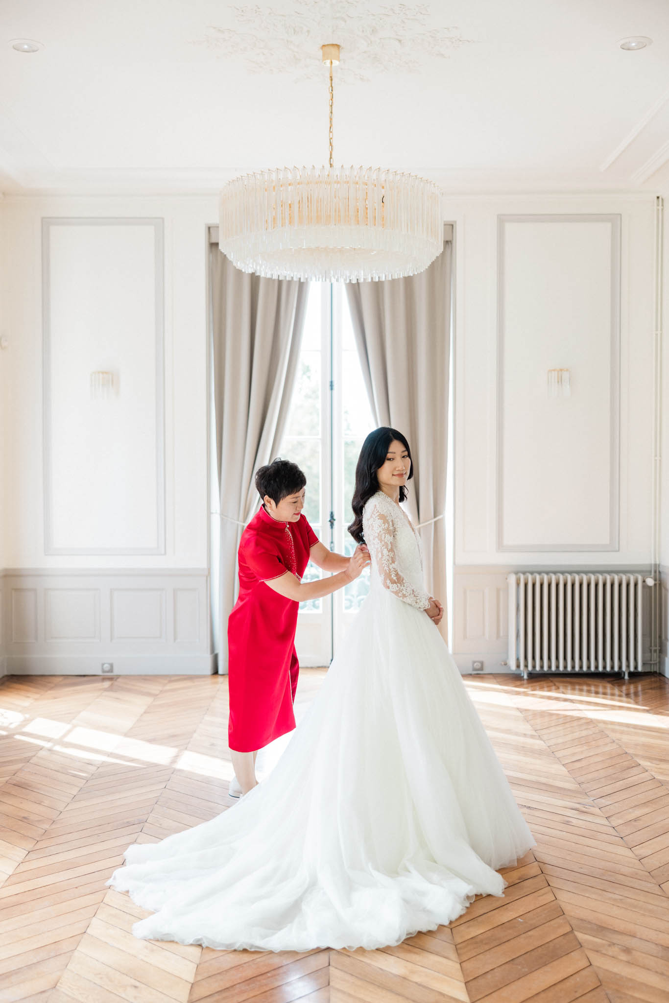 Woman in red cheongsam fastening bride's lace-sleeve gown in white-panelled chateau room with chandelier