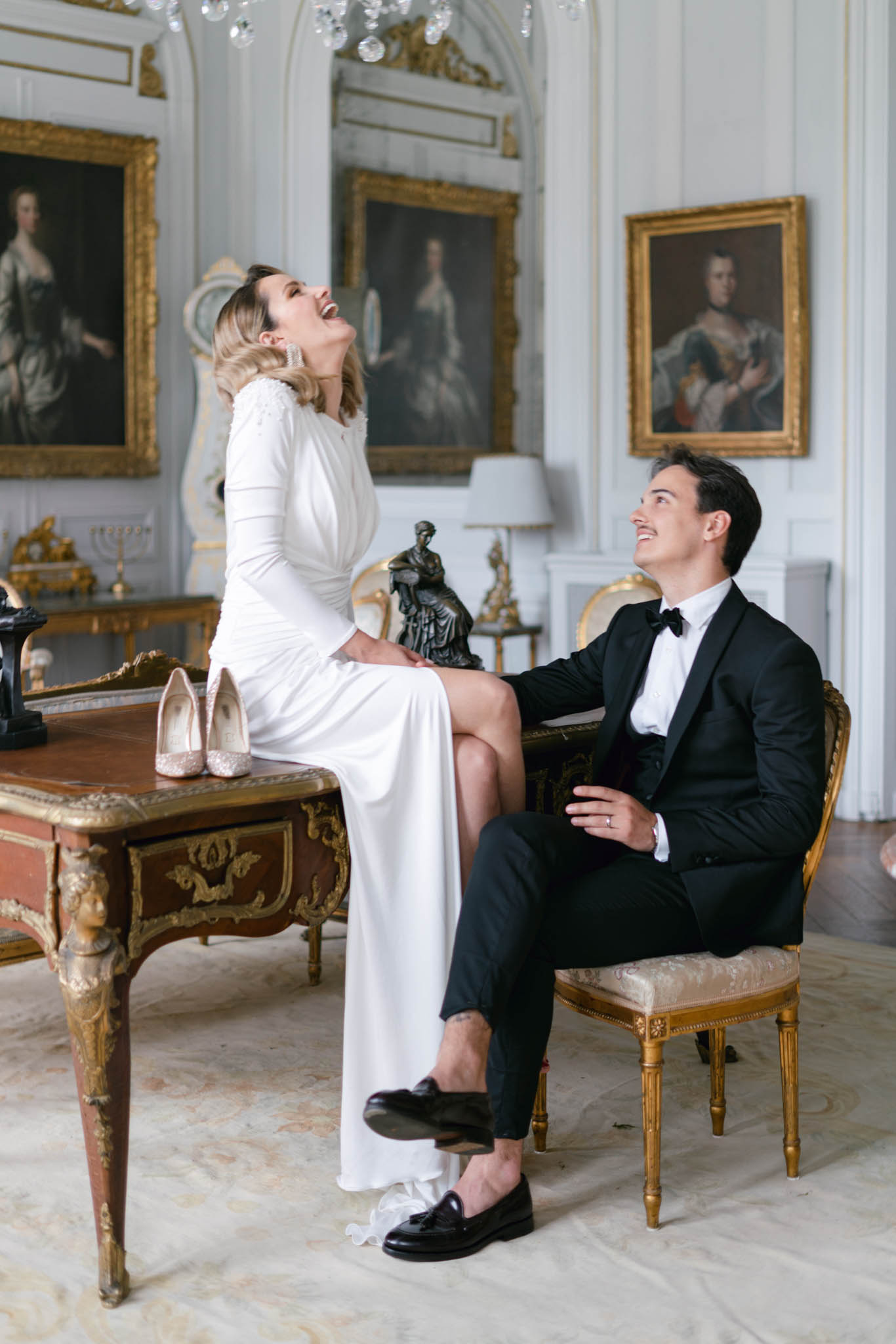 Bride sitting on antique desk laughing with groom in tuxedo inside gilded chateau salon with chandelier