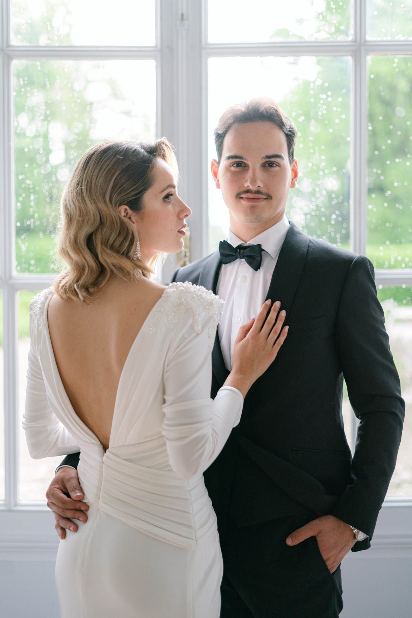 Bride in beaded V-back gown with vintage waves and groom in tuxedo backlit by multi-pane window