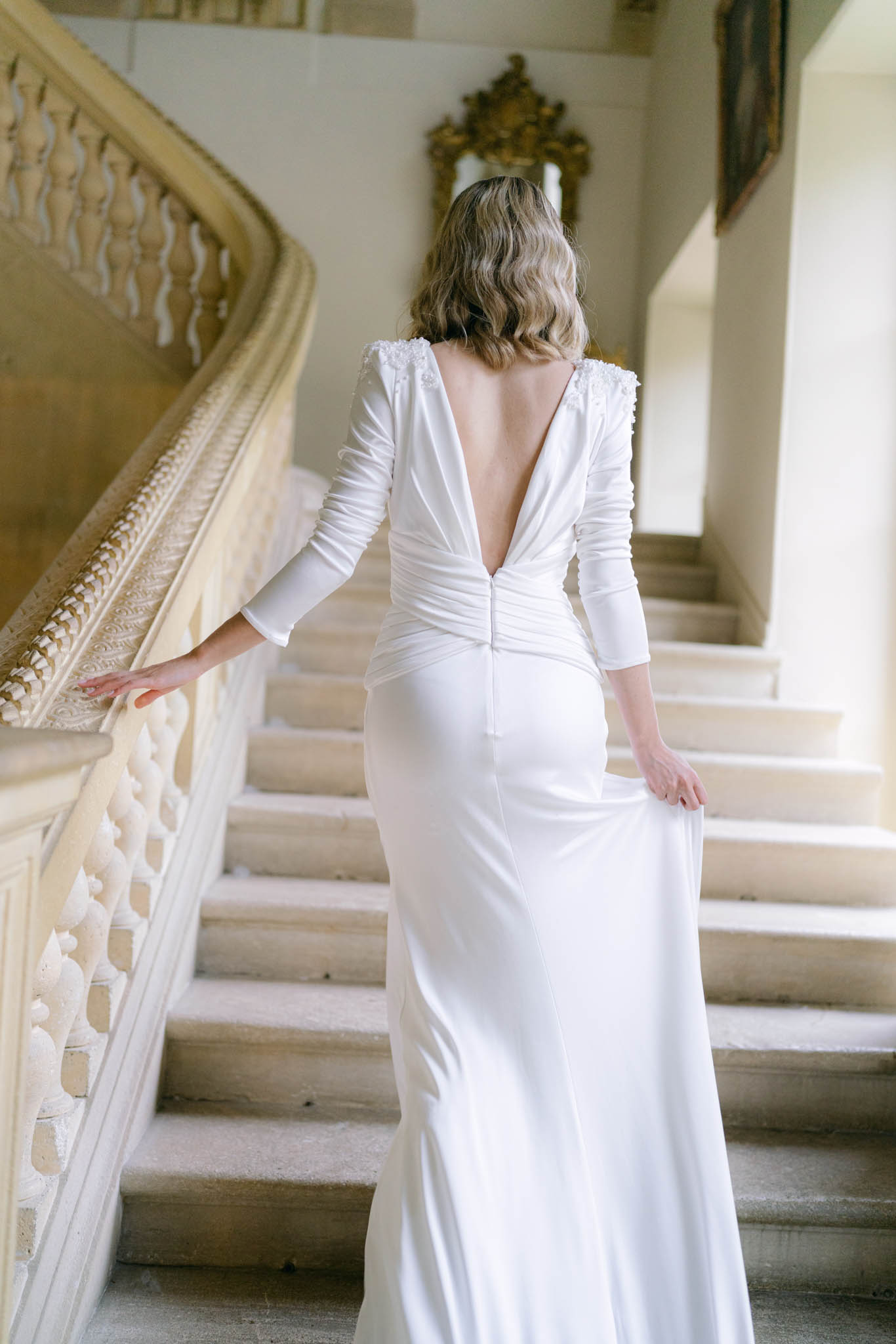 Bride ascending grand staircase from behind in ivory V-back crepe gown with floral applique