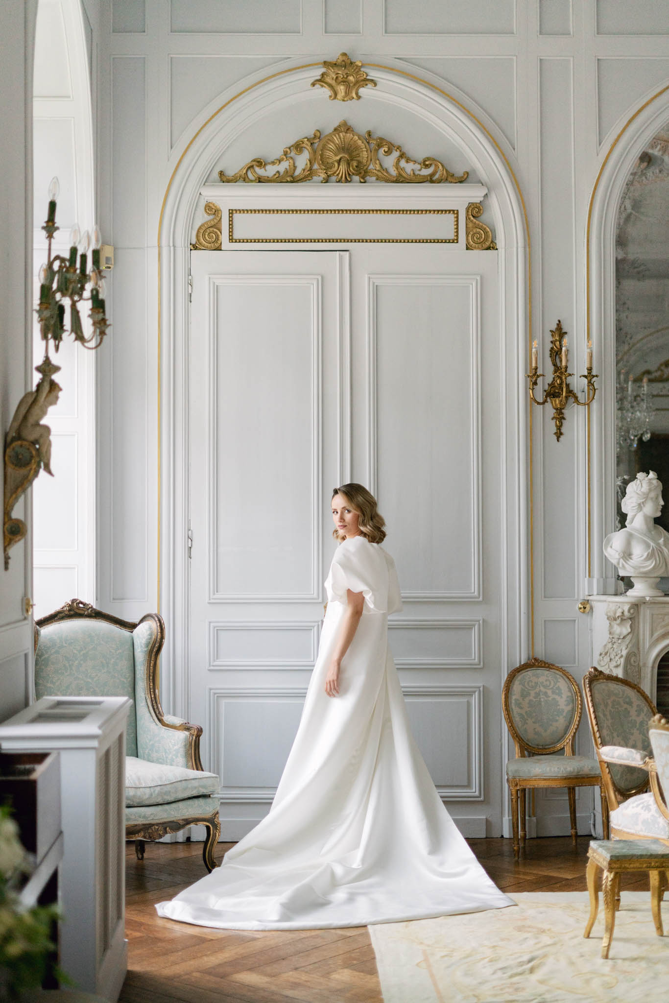 A bridal portrait taken indoors in a French château salon, showing a single bride standing in front of a white-painted boiserie door framed by an arched surround with ornate gilded rococo carvings at the apex. The bride wears an ivory satin A-line gown with flutter sleeves and a long cathedral-length train that spreads across the herringbone parquet floor. She is turned away from the camera with her face looking back over her shoulder. The room features gilded bronze wall sconces with candle-style lights, Louis XVI-style armchairs upholstered in pale mint damask fabric, a white marble fireplace mantel topped with a classical white plaster bust, and a large mirror to the right. The overall interior palette is white, soft grey, and gold, consistent with a classic French grand house aesthetic. Full-length portrait composition. Potential venue feature image.