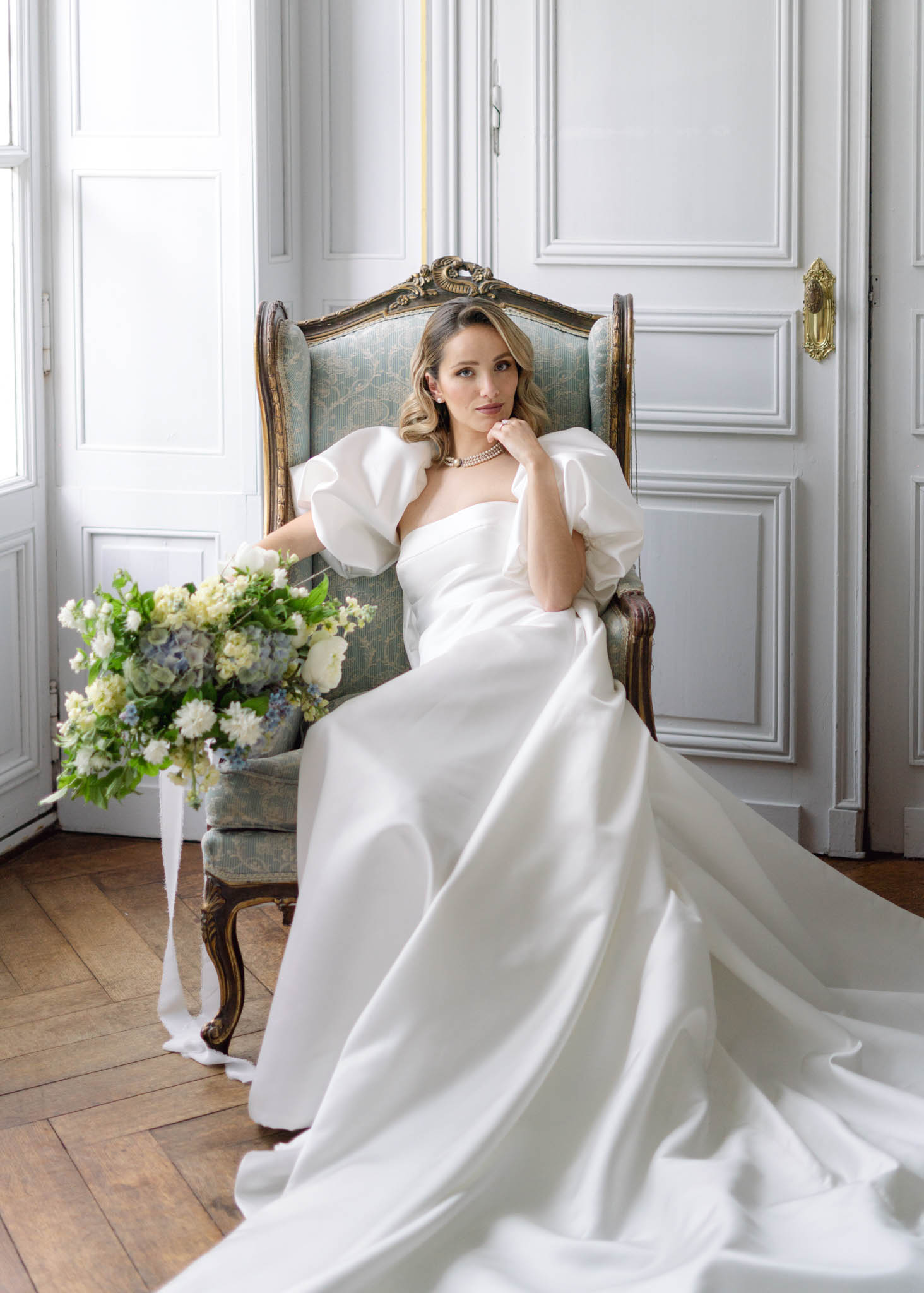 A bridal portrait of a single bride seated in a gilded Louis XV-style wingback armchair upholstered in pale blue-green damask fabric, positioned in an ornate interior room with white wood-panelled walls, gold brass door hardware, and herringbone parquet flooring. The bride wears an ivory structured satin gown with a square neckline and dramatic oversized puff sleeves, paired with a multi-strand pearl choker necklace and white gloves; the full skirt and long train pool across the floor. She holds a loose, garden-style bouquet resting against the chair arm, composed of soft white blooms, pale blue hydrangeas, delicate blue forget-me-nots, and lush green foliage with long white ribbon streamers trailing to the floor. The overall styling is classic French interior with a modern bridal silhouette. Medium portrait shot, framed from floor to ceiling, natural window light coming from the left.