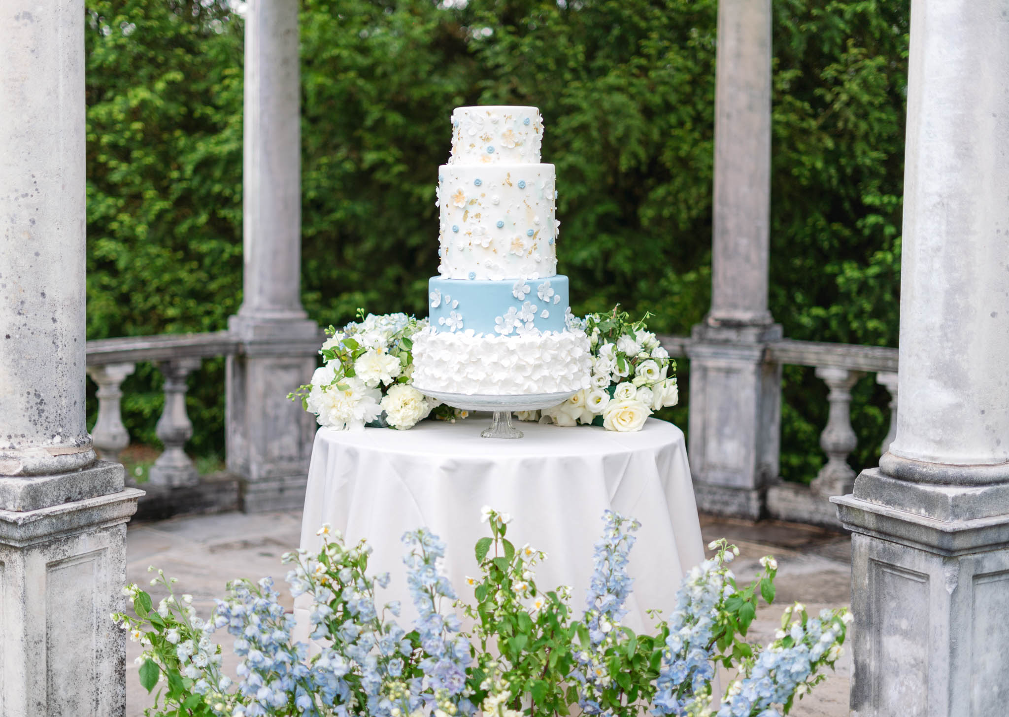 Four-tier powder-blue and white cake with sugar flowers and gold leaf beside delphinium on stone colonnade