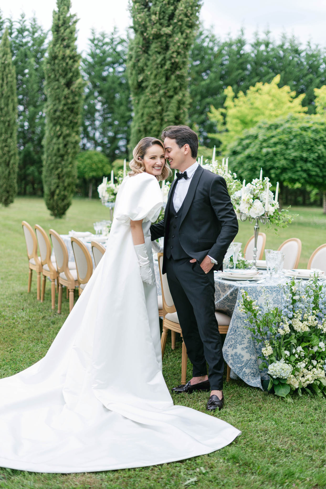 Bride and groom touching foreheads beside a blue toile tablecloth reception table with white hydrangeas and silver candela...