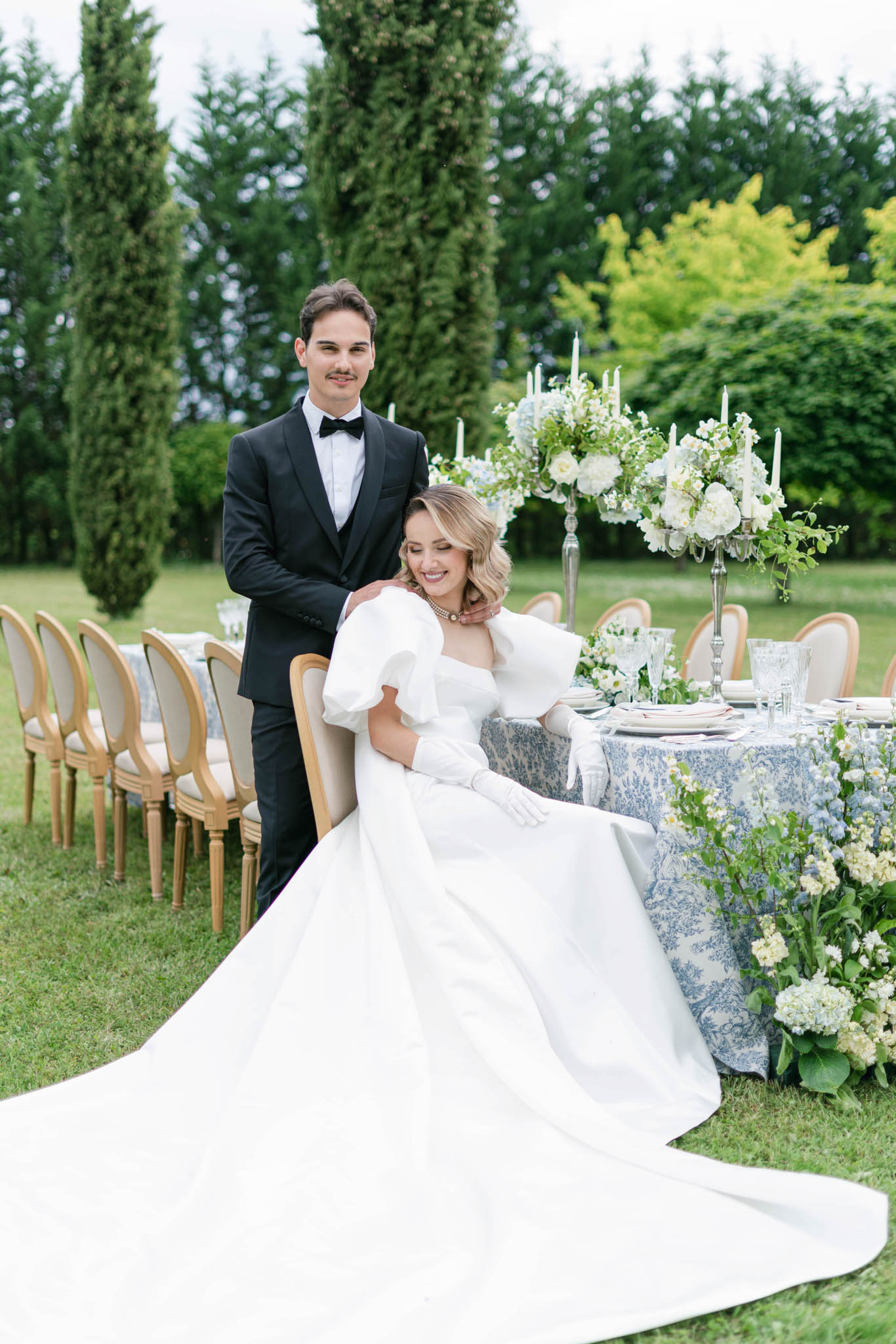 A couple poses together at an outdoor garden reception tablescape, with the bride seated and the groom standing behind her with his hand on her shoulder. The bride wears a white ball gown with dramatic oversized puff sleeves, long white gloves, and a pearl necklace, with her blonde hair styled in soft waves; the groom wears a black tuxedo with a bow tie. The reception table is dressed in a blue and white toile de Jouy linen, set with crystal glassware, stacked plates, and silver candelabras holding tall white taper candles. Tall centerpiece arrangements of white hydrangeas, white roses, and trailing greenery sit on silver stands, with additional white hydrangea and delphinium clusters arranged at the base of the table. Gold Louis XVI-style chairs line the length of the table behind them. The overall styling palette is white, ivory, and dusty blue with a classic French aesthetic. This is a medium portrait shot taken outdoors on a garden lawn flanked by tall cypress trees.