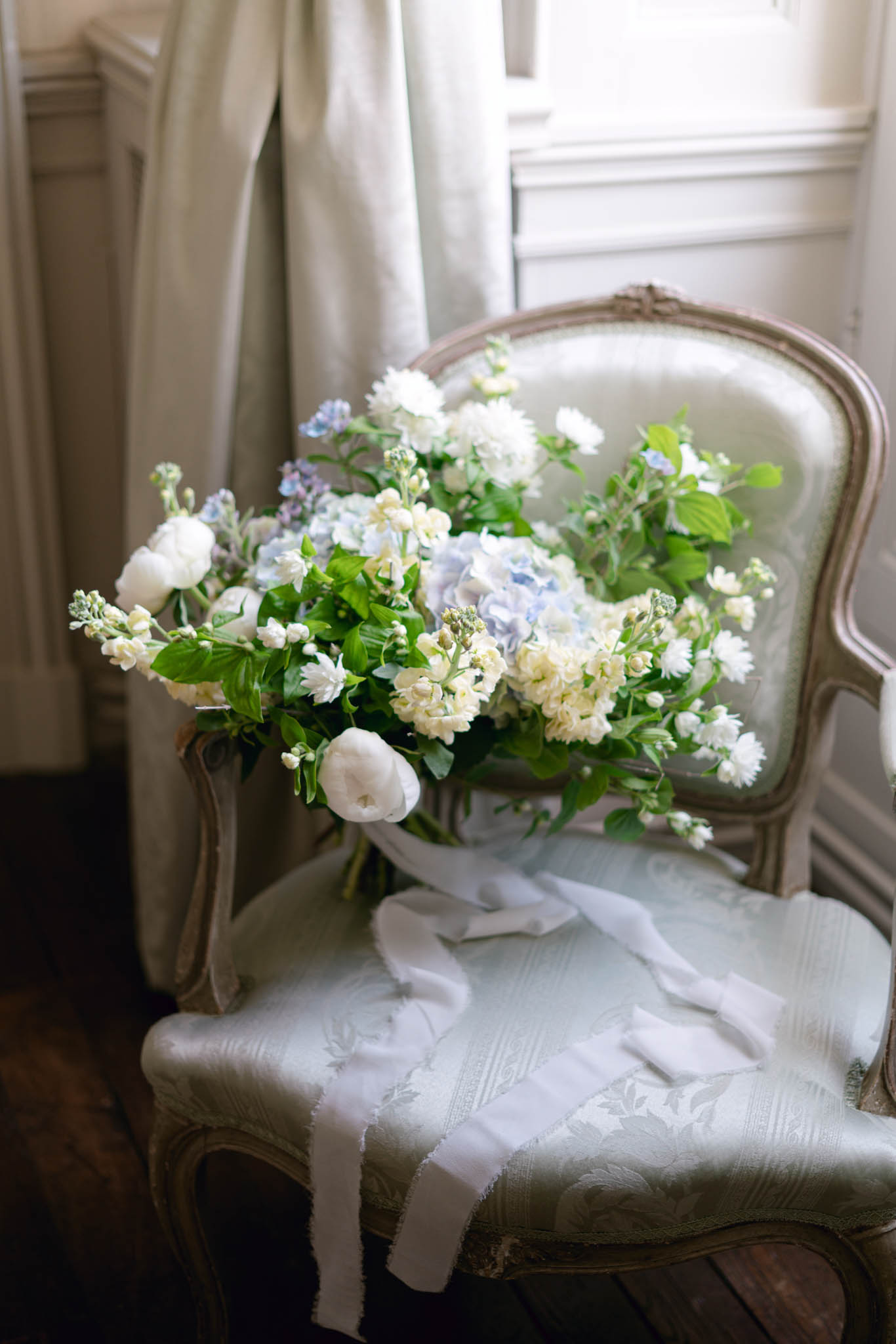 Bridal bouquet of white ranunculus, pale blue hydrangeas, and cream stock on mint-green damask bergere chair