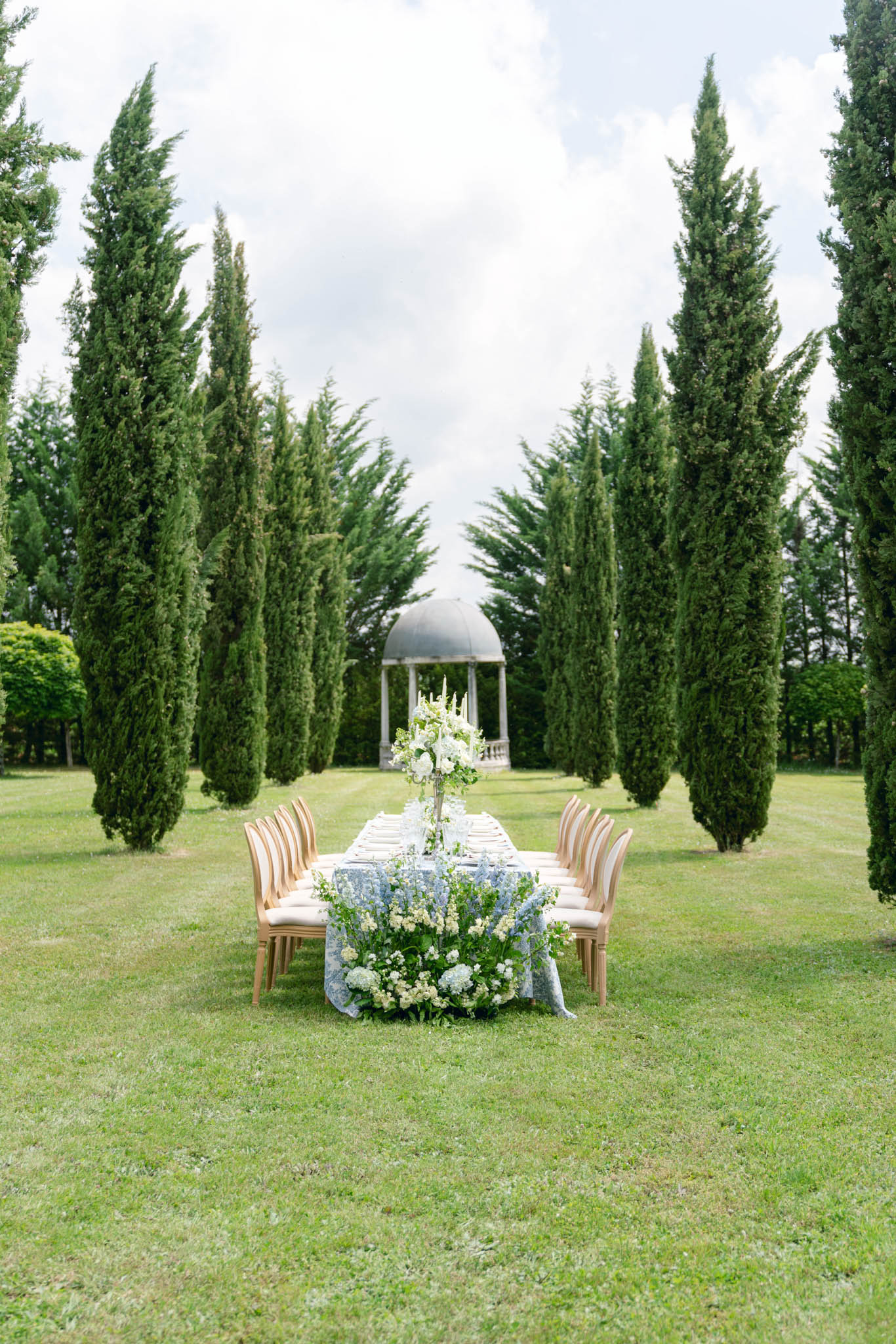 An outdoor reception tablescape styled in a formal garden setting, with no guests present. A long rectangular dining table is set along a central grass allée flanked by tall Italian cypress trees, with a classical stone domed rotunda/gazebo visible at the far end of the avenue. The table is dressed with a pale blue toile or floral-print linen tablecloth and set with white plates, glassware, and place settings for approximately 12 guests. A tall, slender candelabra-style floral centerpiece rises from the center of the table, topped with white blooms and taper candles, while the base of the table is heavily garlanded with a lush ground-level arrangement of white hydrangeas, white stock flowers, pale blue delphiniums, and green foliage spilling across the linen. The chairs are natural wood French Louis-style with cream upholstered seats. The shot is a wide, symmetrical perspective taken from the end of the table looking toward the rotunda, emphasizing the formal axis of the garden. Potential venue feature image.