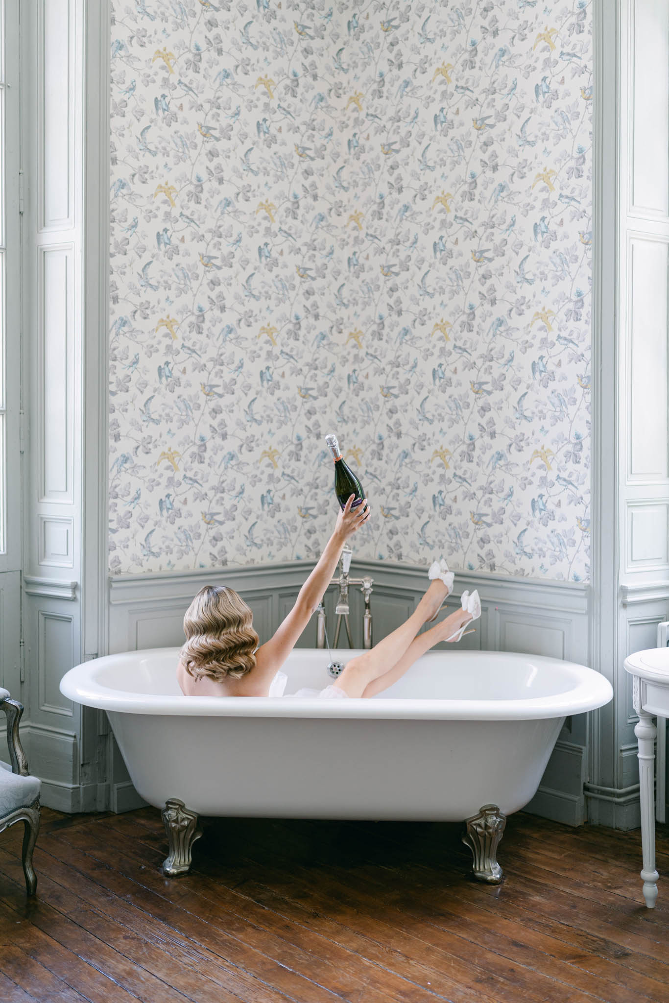 Bride poses in clawfoot tub holding champagne bottle in bird-print wallpapered chateau room from behind