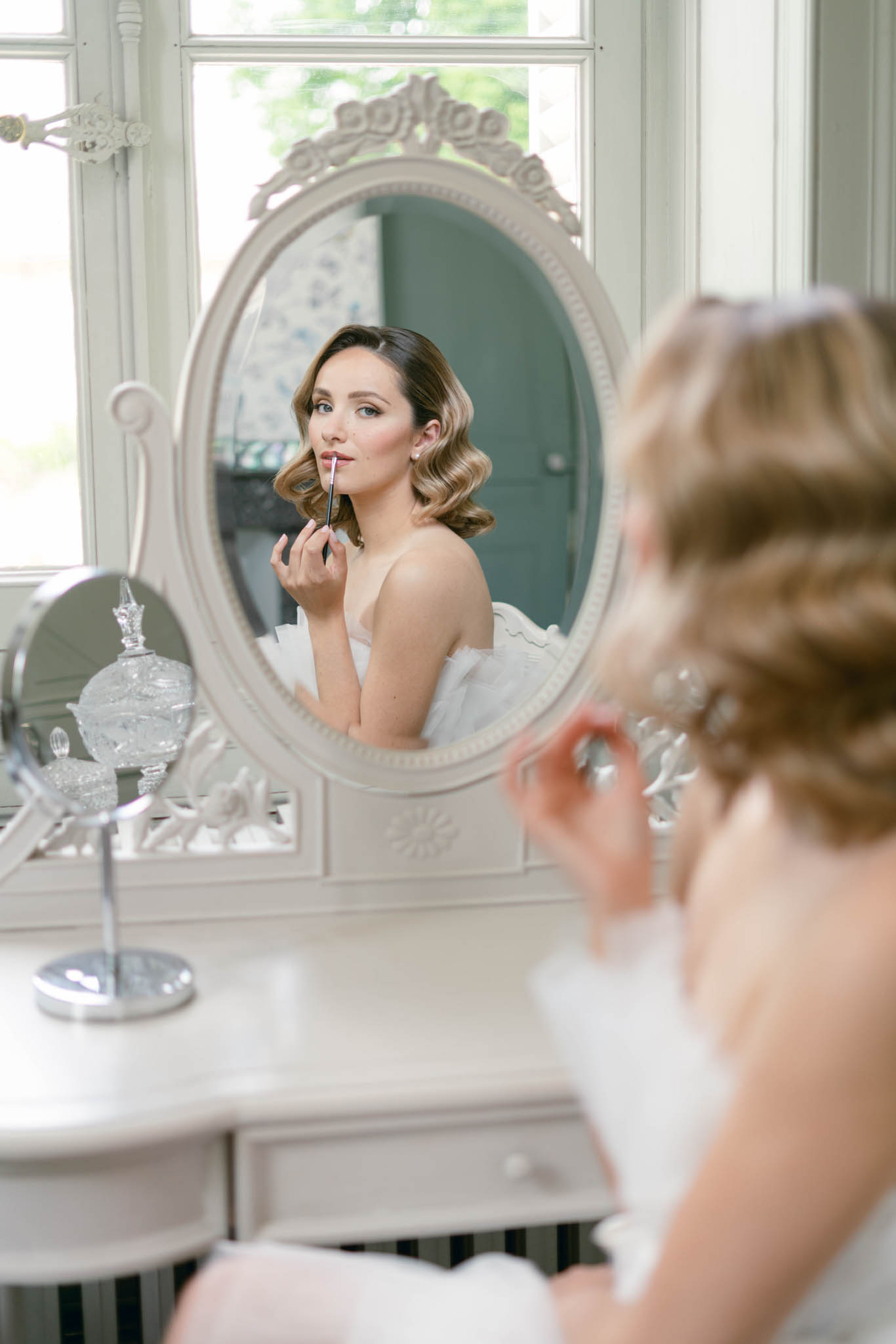 Bride applying lip color at ornate dressing table reflected in oval mirror with vintage waves hairstyle