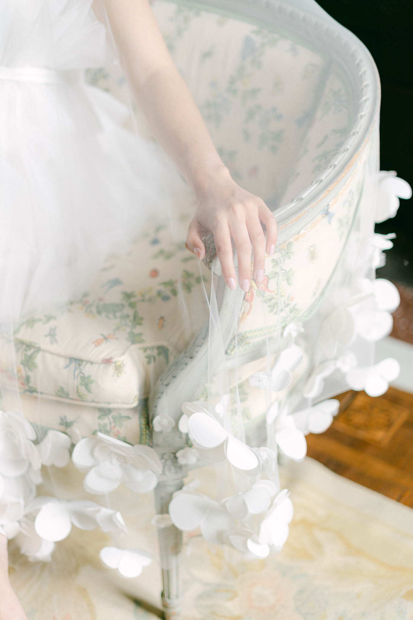Close-up of bride's hand on floral toile armchair showing white tulle gown with petal applique hem detail