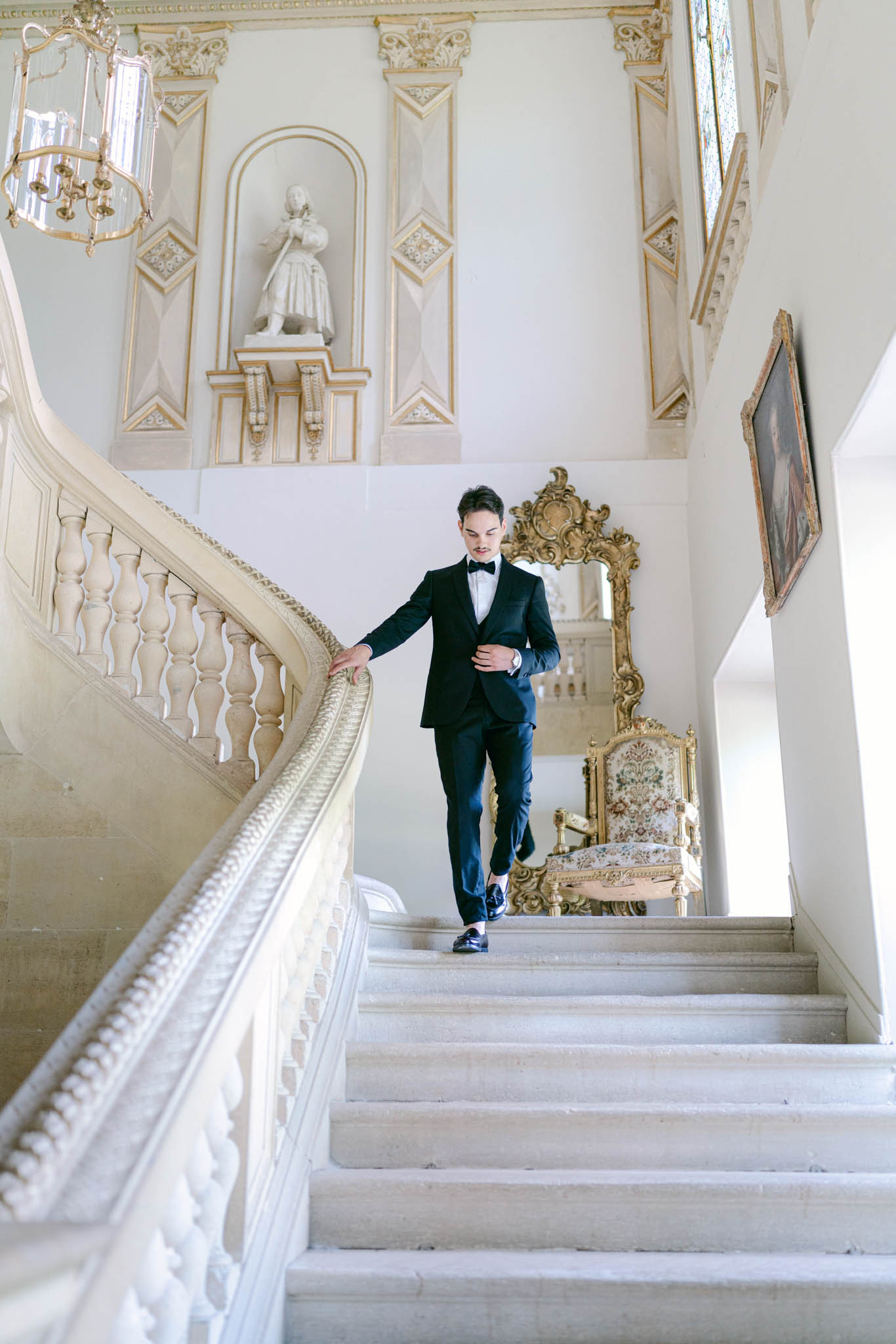 Groom in navy tuxedo descending a grand staircase inside an ornate French chateau