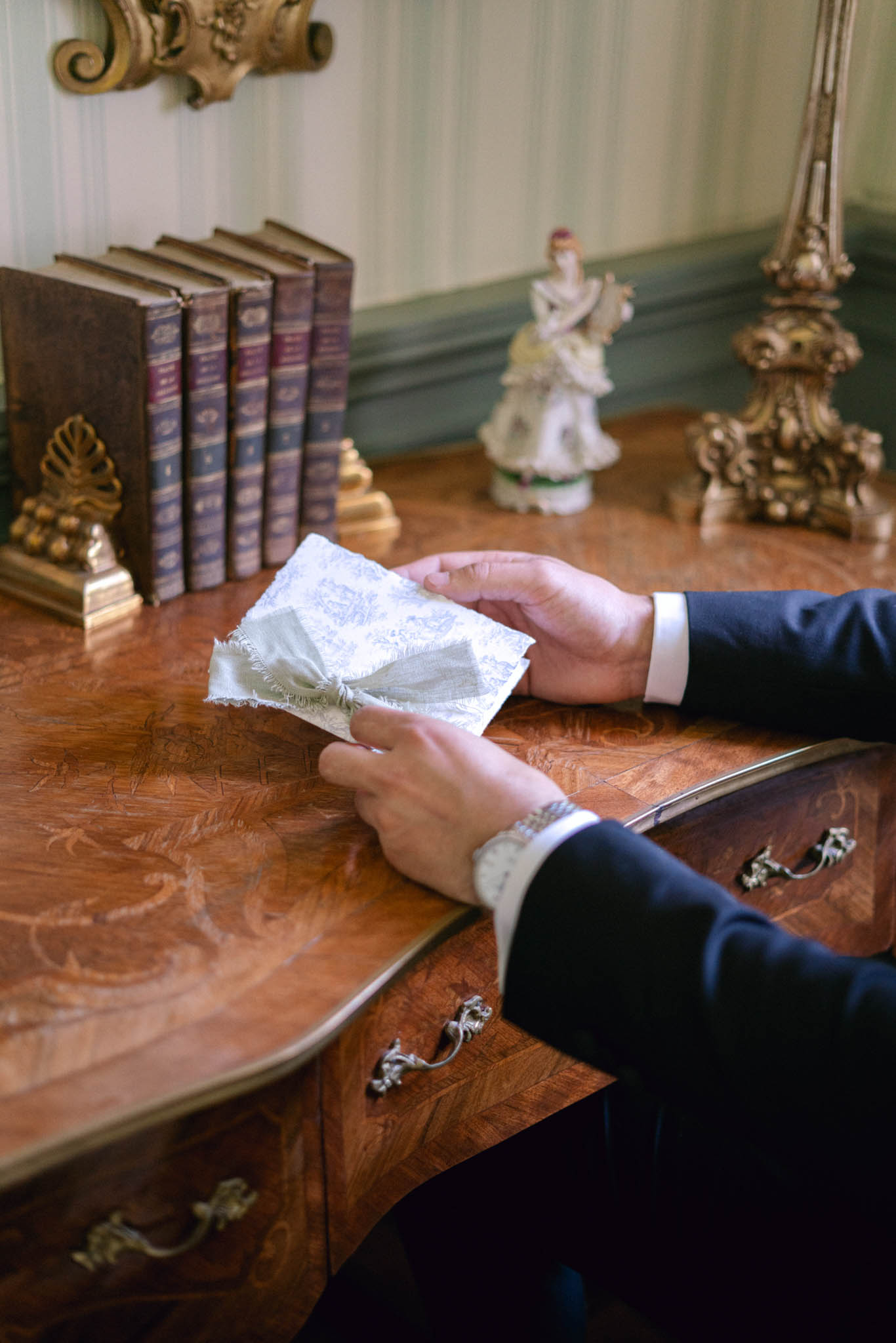 Groom's hands holding toile de Jouy handkerchief tied with grey ribbon at antique desk in French interior
