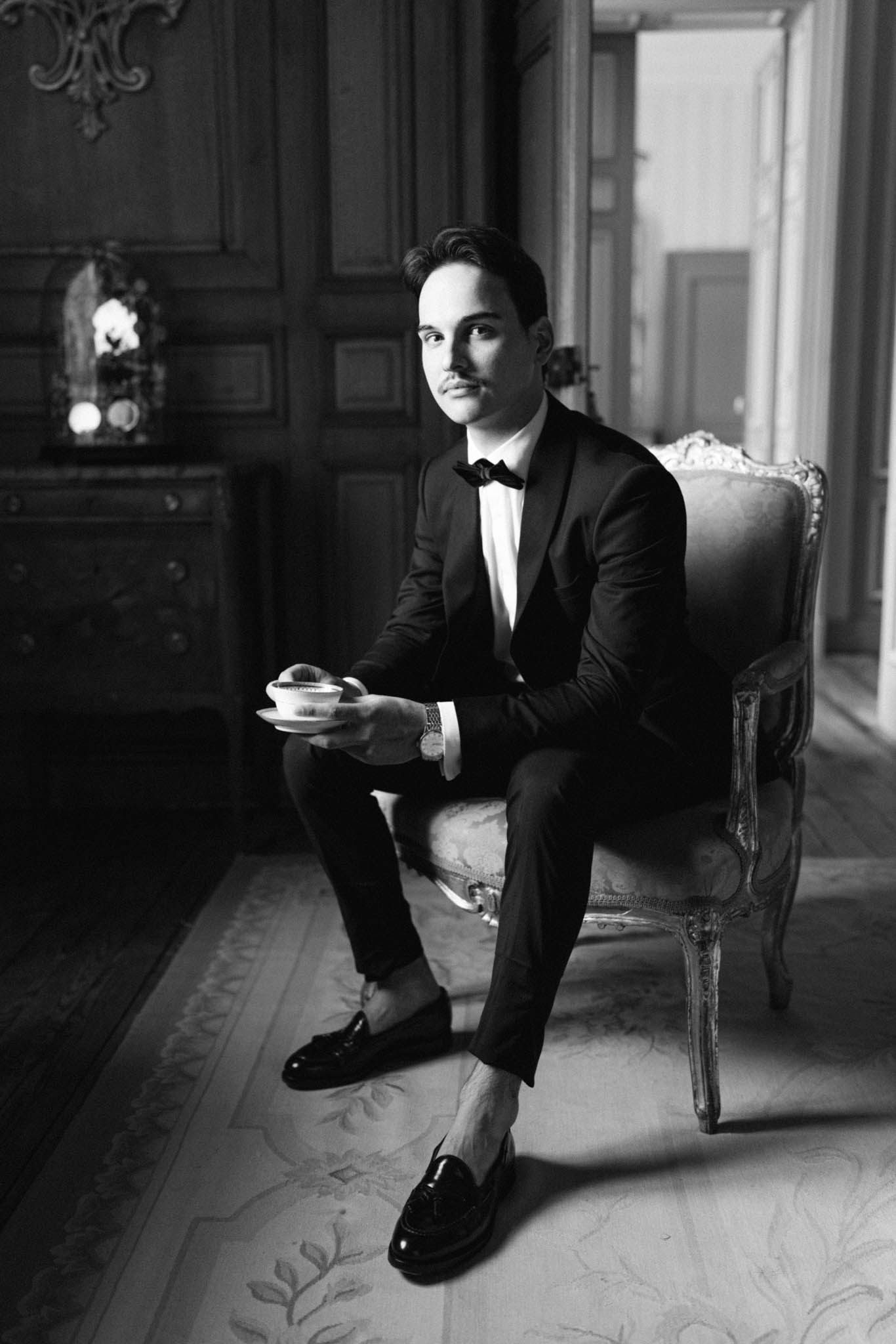 Black and white portrait of groom in tuxedo holding teacup in carved Louis XVI armchair