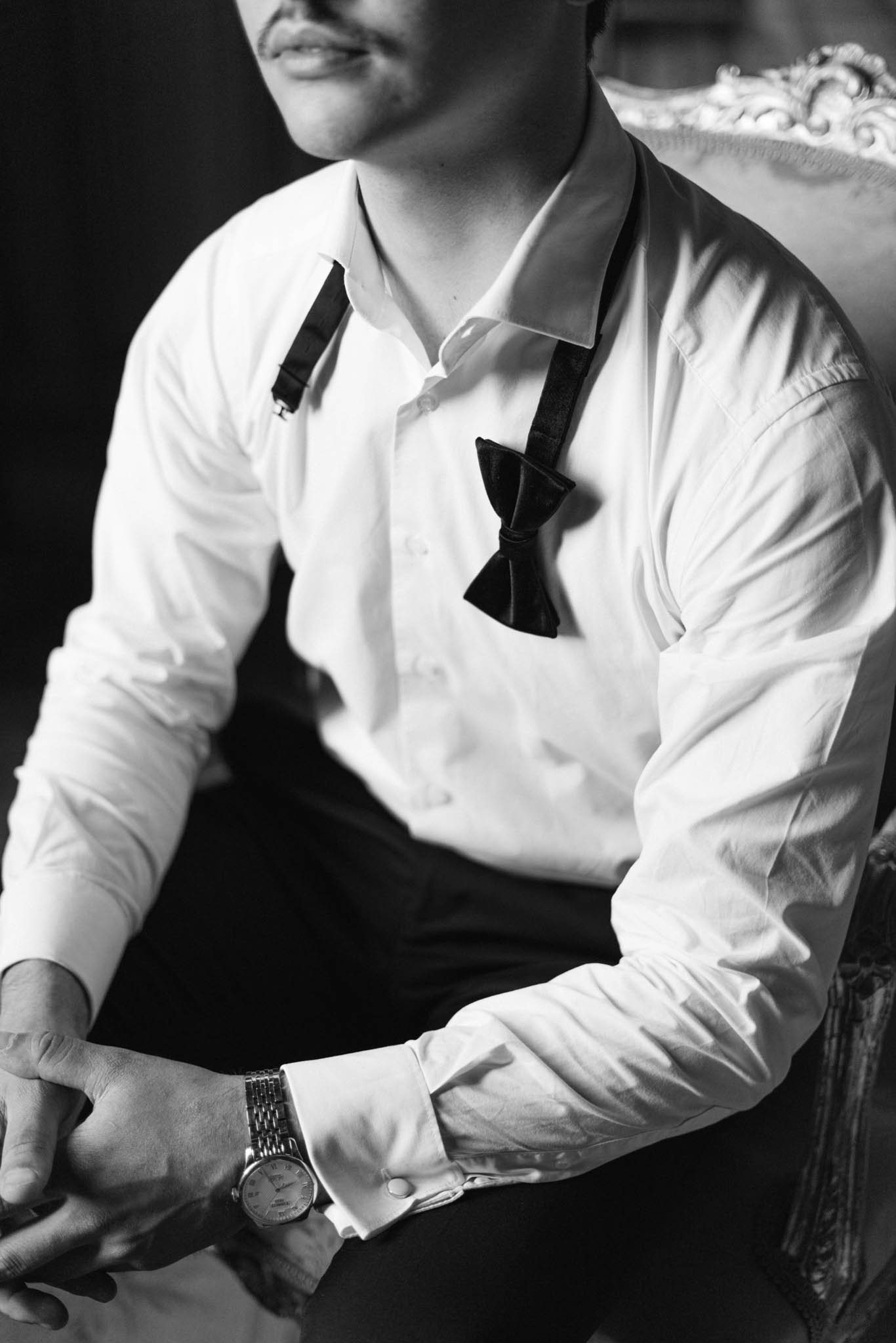 This black-and-white close-up portrait captures a groom during the getting-ready phase, seated in what appears to be an ornately carved chair visible at the upper right edge of the frame. He is wearing a white dress shirt with French cuffs, dark suspenders, and an undone black bow tie hanging loosely around his neck, suggesting he is mid-preparation. A classic metal-bracelet dress watch is visible on his left wrist, and his hands are loosely clasped in his lap. The image uses strong contrast with a dark background that draws focus to the white shirt and accessories, shot in a tight portrait composition that crops just below the lips and above the knees.