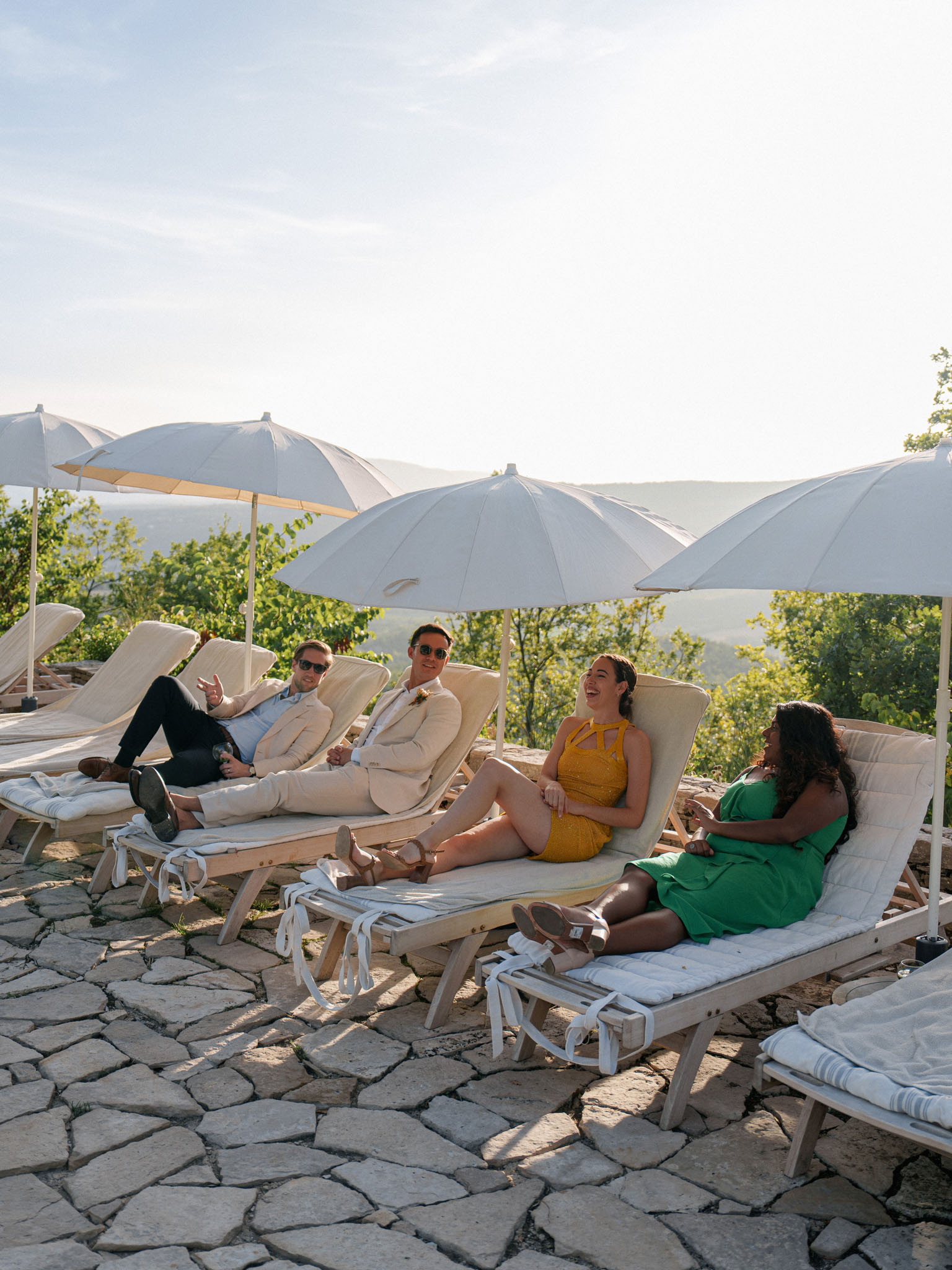 Four wedding guests relaxing on wooden sun loungers under white umbrellas overlooking wooded hillside