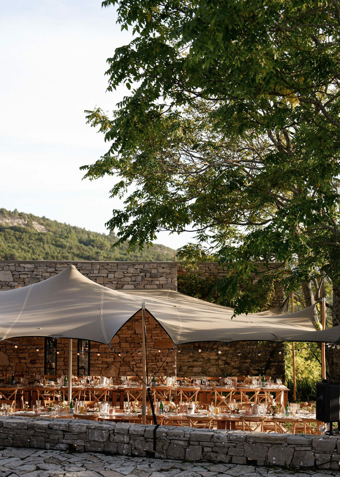An outdoor wedding reception setup on a stone-paved terrace surrounded by rustic stone walls, with long wooden banquet tables and wooden folding chairs arranged beneath ivory stretch tents. The tables are set with glassware, green glass wine bottles, candles, and low floral centerpieces in warm, muted tones. String lights are strung between the tent poles, adding soft ambient lighting. The overall styling is relaxed and rustic, with natural wood tones and neutral textile choices. This is a wide establishing shot showing the full reception layout with no guests present. Potential venue feature image.