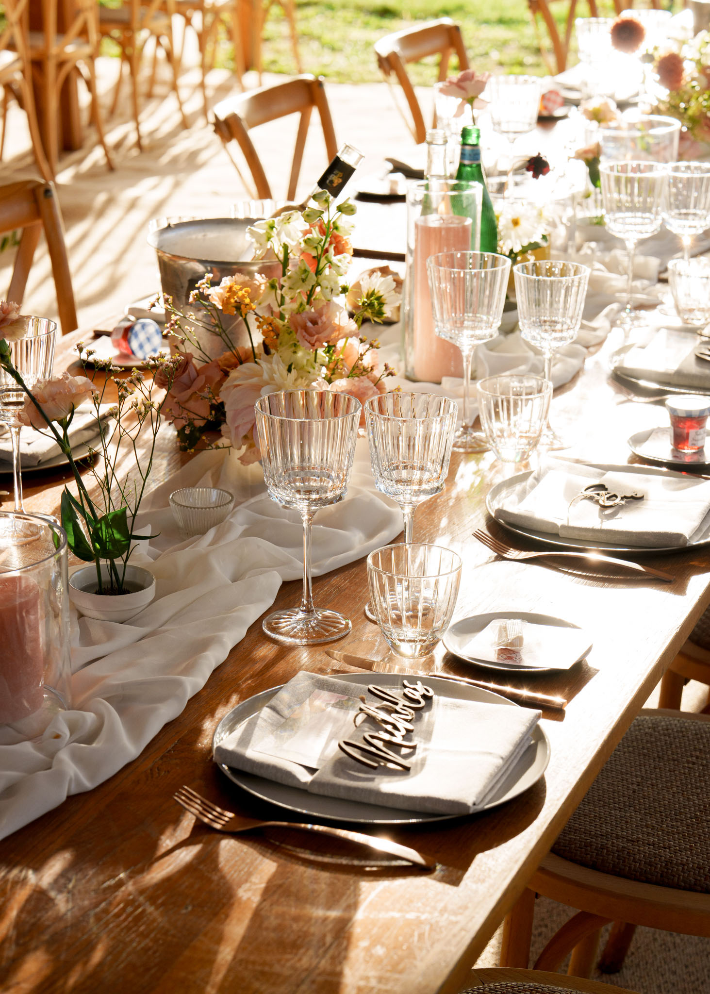 Farm table with blush ranunculus, peach carnations, laser-cut name cards, grey chargers, and bronze flatware