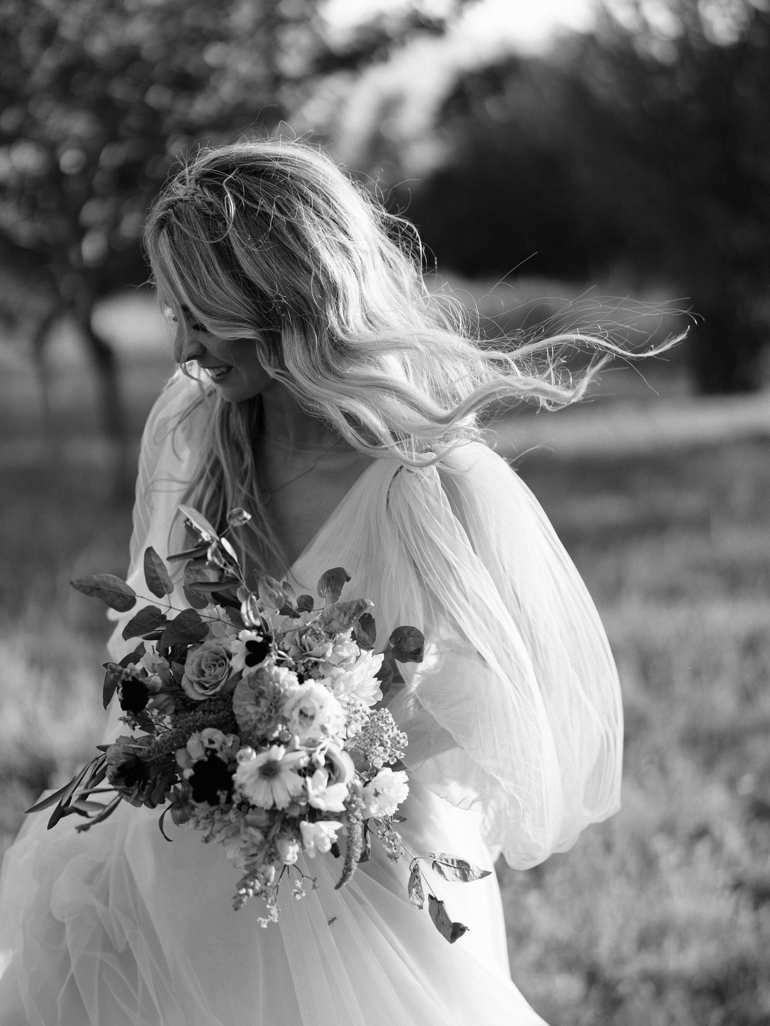 This is a black-and-white outdoor bridal portrait of a single bride photographed in three-quarter frame against a softly blurred outdoor backdrop of trees. The bride's long, wavy blonde hair is caught mid-movement by the wind, sweeping across her shoulder as she looks downward with a smile. She wears a flowing wedding dress with voluminous, billowy sleeves in a lightweight fabric that also lifts in the breeze. She holds a large, loosely arranged bouquet featuring a mix of roses, dahlias, anemones, daisies, and ranunculus with eucalyptus and mixed foliage throughout; in the monochrome tones, the blooms range from deep darks to bright whites with strong contrast. The image is shot with a shallow depth of field, rendering the background fully out of focus and drawing attention to the movement of the hair and dress. The high-contrast black-and-white processing emphasizes texture in the fabric and floral details.