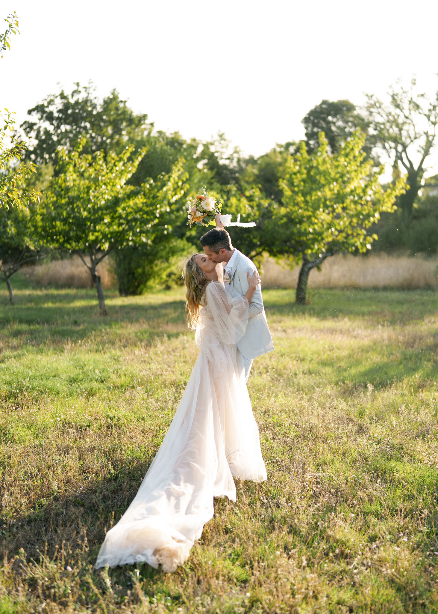Couple kissing in orchard bride raising peach and white bouquet wearing ivory bell-sleeve boho gown