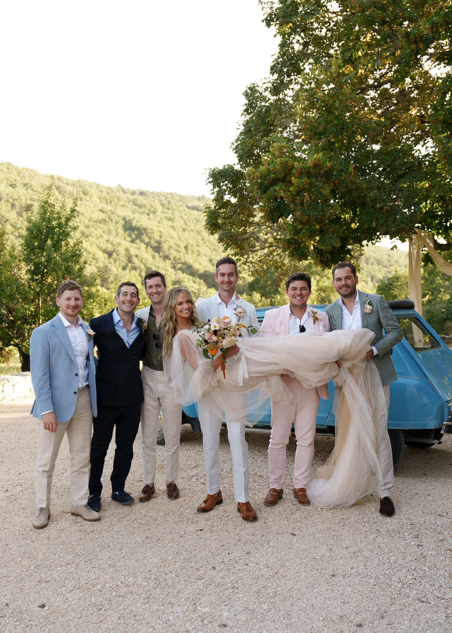 Groom in white suit holds bride while five groomsmen pose by vintage teal Citroen 2CV on gravel driveway