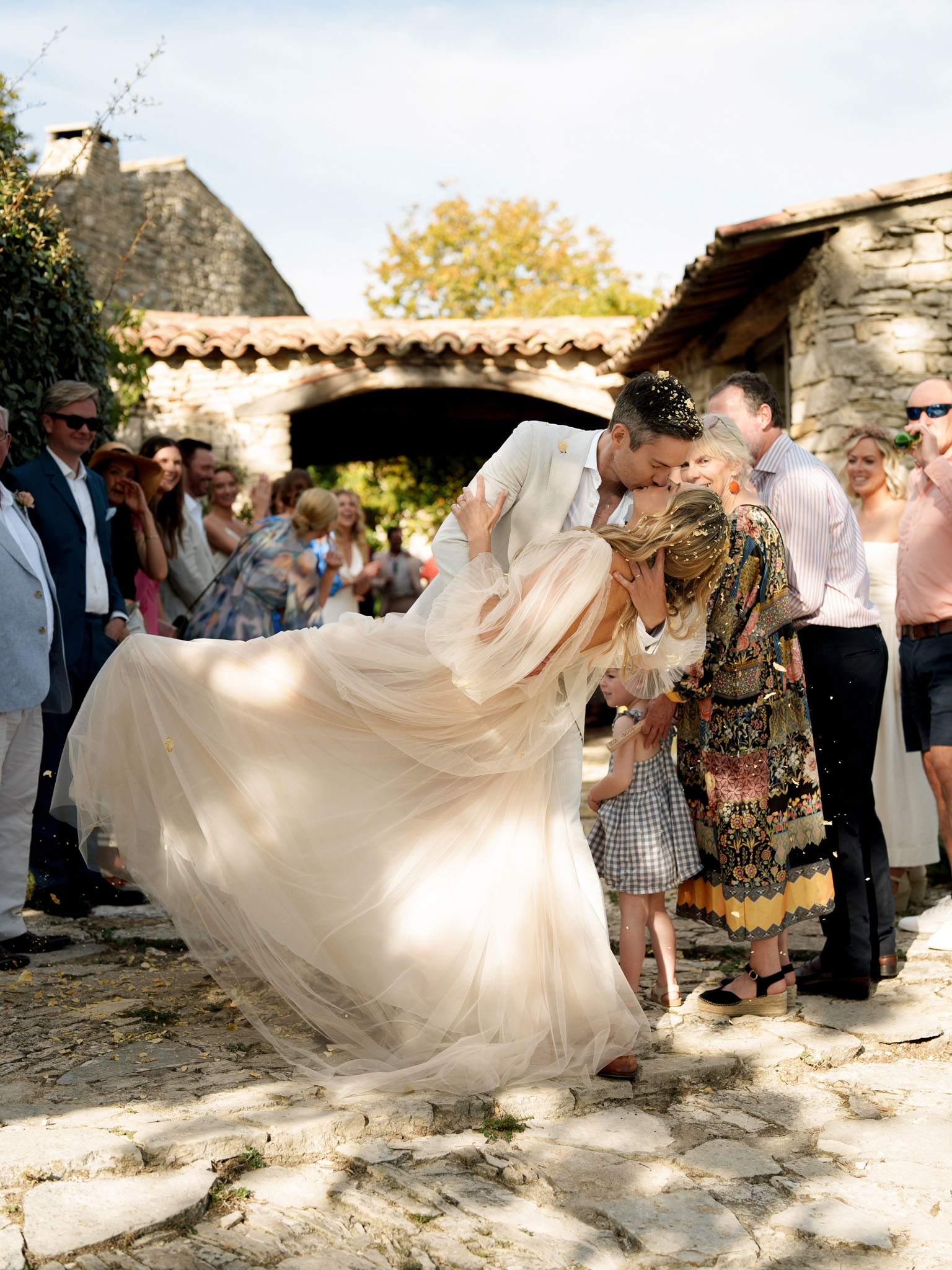 Bride and groom dip kiss with gold confetti as guests cheer on Provencal cobblestone courtyard