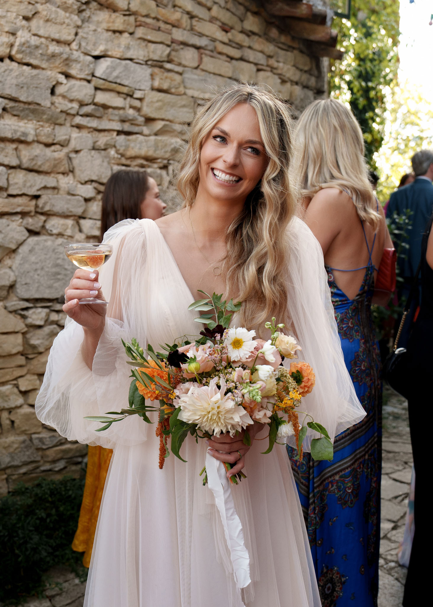 Bride in blush pink sheer-sleeve dress holding garden bouquet of dahlias, peonies, and orange ranunculus