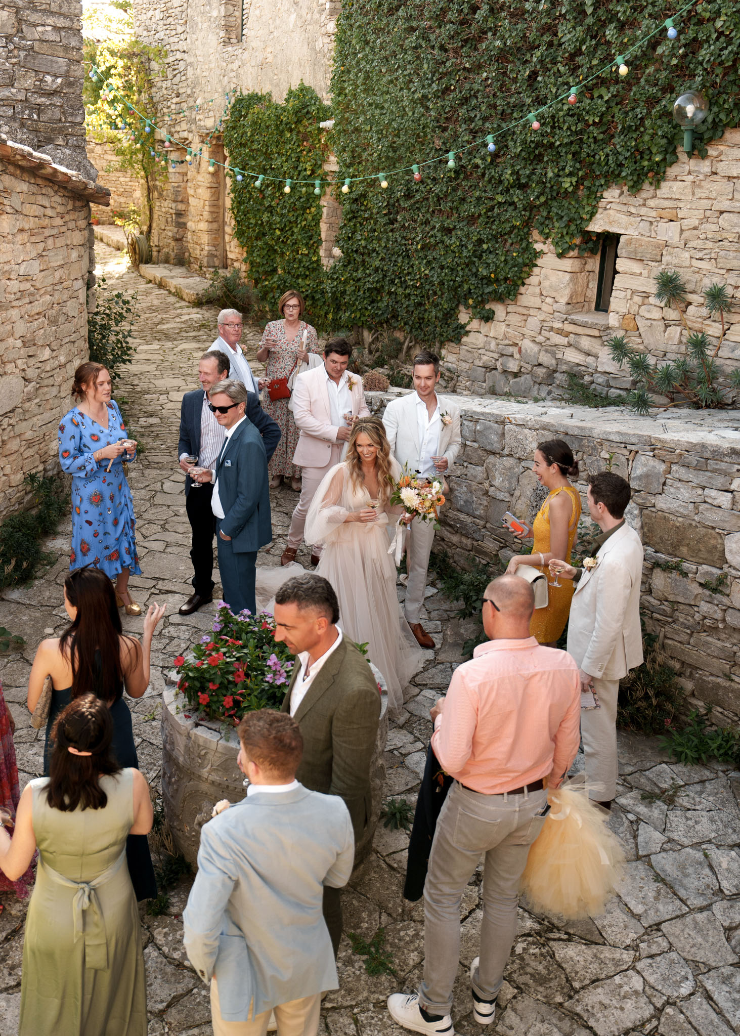 A cocktail hour scene photographed from an elevated angle, showing approximately 15 guests and the bride mingling in a narrow stone-paved courtyard or alleyway flanked by rustic limestone walls covered in climbing ivy. The bride wears a floor-length blush tulle gown with sheer long sleeves and a floral crown, holding a loose bouquet of peach, white, and orange blooms including dahlias and greenery. Guests are dressed in a mix of colorful and neutral outfits including a blue floral midi dress, a mustard yellow sleeveless dress, an olive green suit, a blush pink blazer, and various linen-toned suits, giving the gathering a relaxed, boho-rustic feel. A string of colorful festoon bulb lights is strung between the stone walls overhead, adding a festive decorative touch to the outdoor space.
