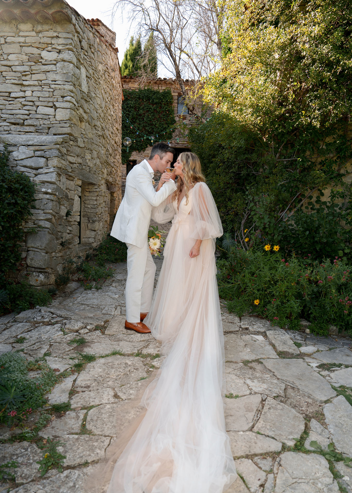 A couple shares a champagne toast in an outdoor courtyard of a rustic Provençal stone property, leaning in close as if about to kiss over the glasses. The groom wears a cream linen suit with tan loafers, and the bride wears a blush pink tulle gown with voluminous sheer cape sleeves and a long flowing train that pools across the flagstone paving. The setting is a narrow stone-paved alleyway between old limestone buildings with globe string lights visible in the background archway. The styling is relaxed boho with warm, natural tones, and a cluster of yellow and orange flowers is visible mid-ground. The shot is a full-length portrait taken at a slight distance, capturing the full train of the dress and the surrounding courtyard garden.