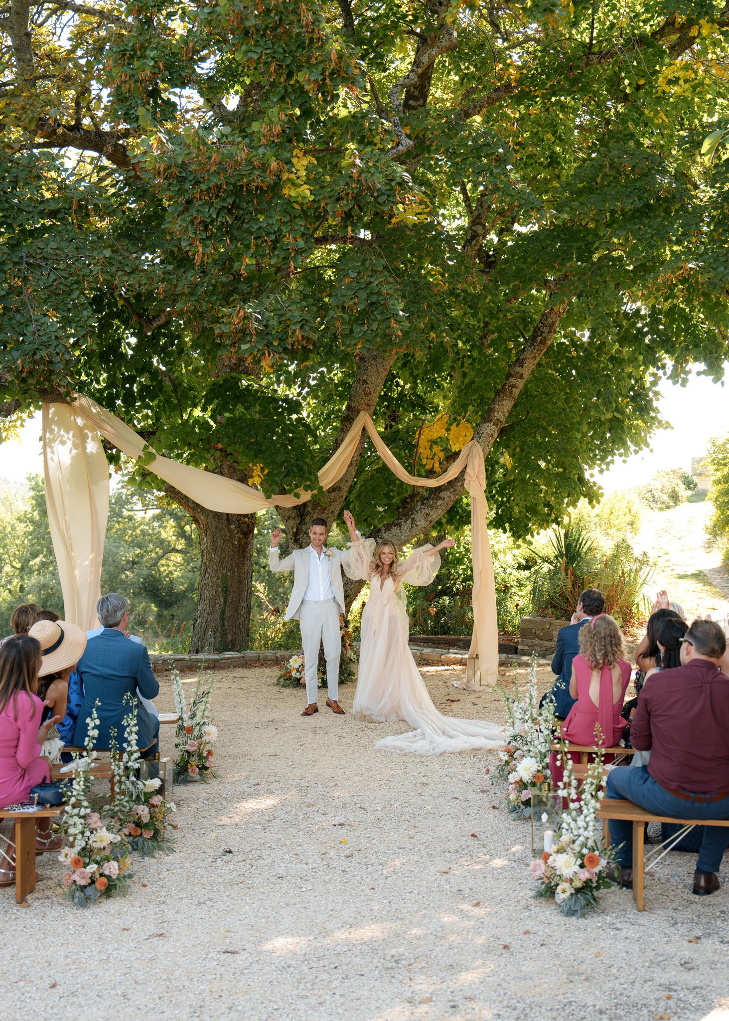 Couple raises hands in recessional under fabric canopy with peach and coral aisle flowers on gravel